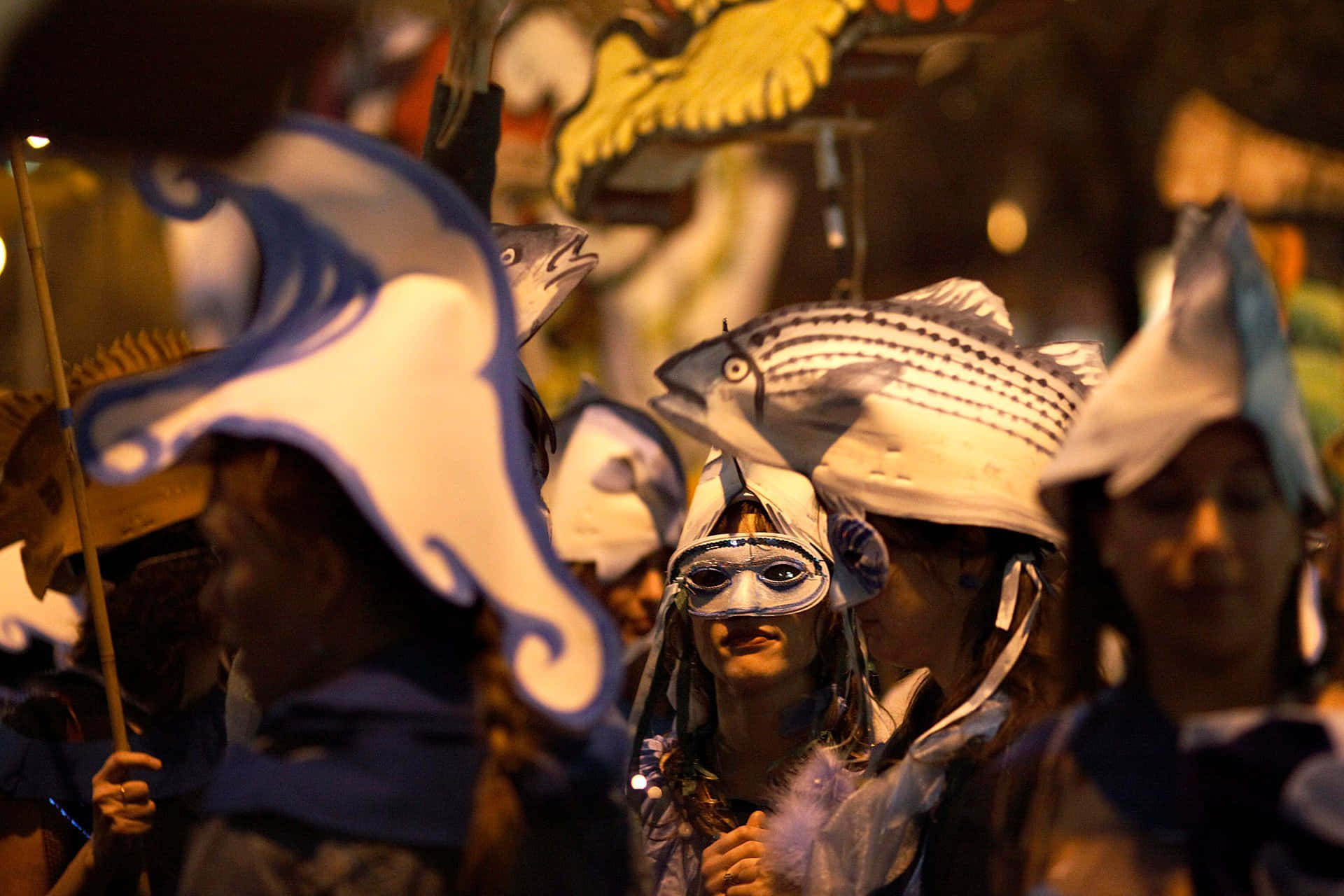 Halloween Parades 桌布 壁紙 — 人們戴著魚主題面具和服裝，在夜晚遊行，背景有溫暖發光的燈光。