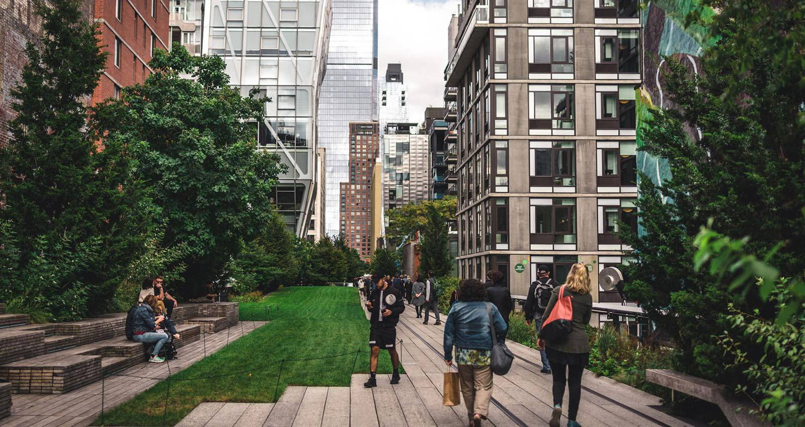 New York 桌布 壁紙 — 人們沿著一條充滿活力的綠色城市公園小徑漫步和放鬆，小徑兩旁是高聳的現代建築和茂密的樹木。