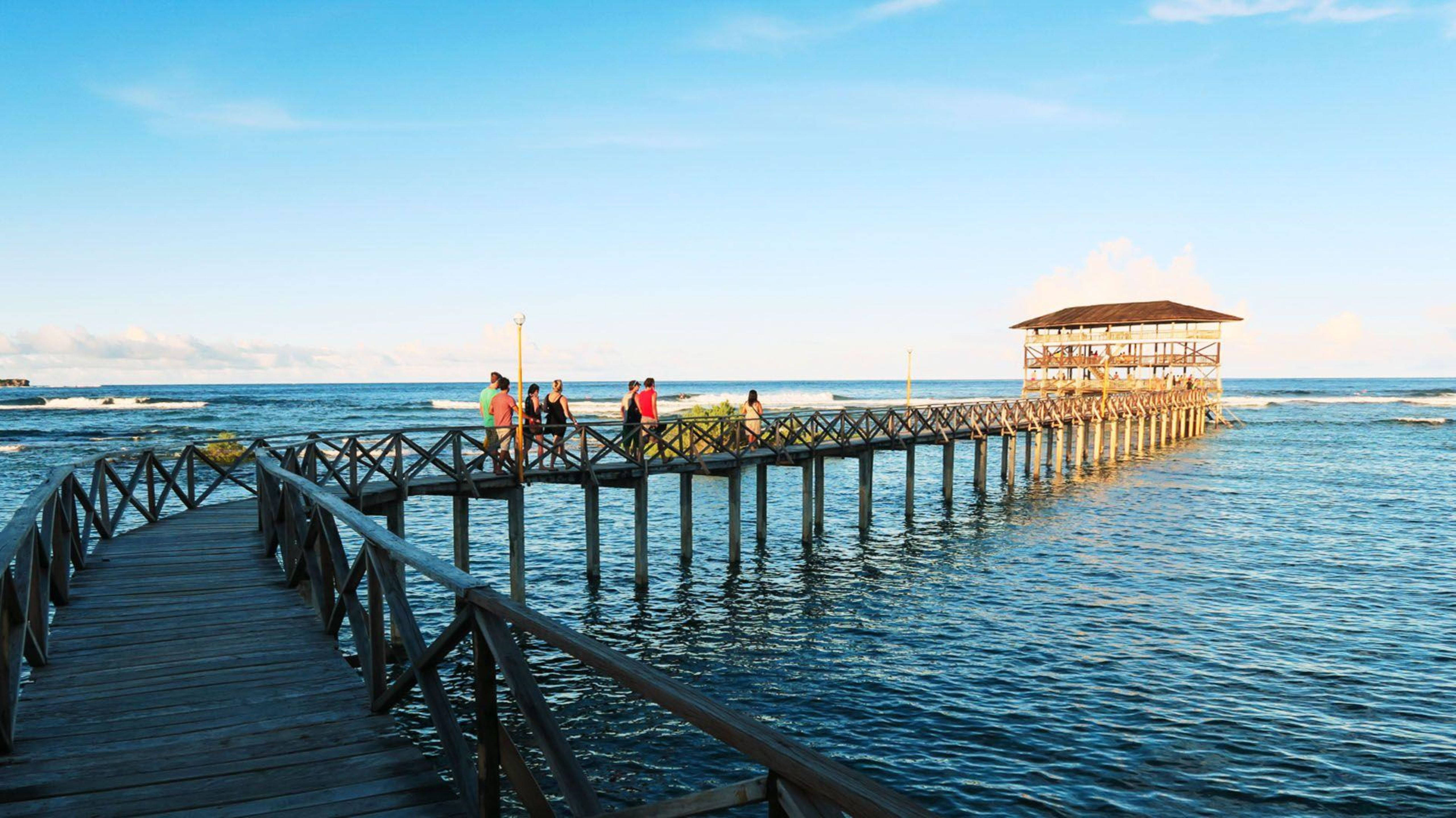 Siargao 桌布 壁紙 — 人們沿著一條長長的木碼頭漫步，在晴朗的天空下，在黃金時段延伸至平靜的藍色海洋。