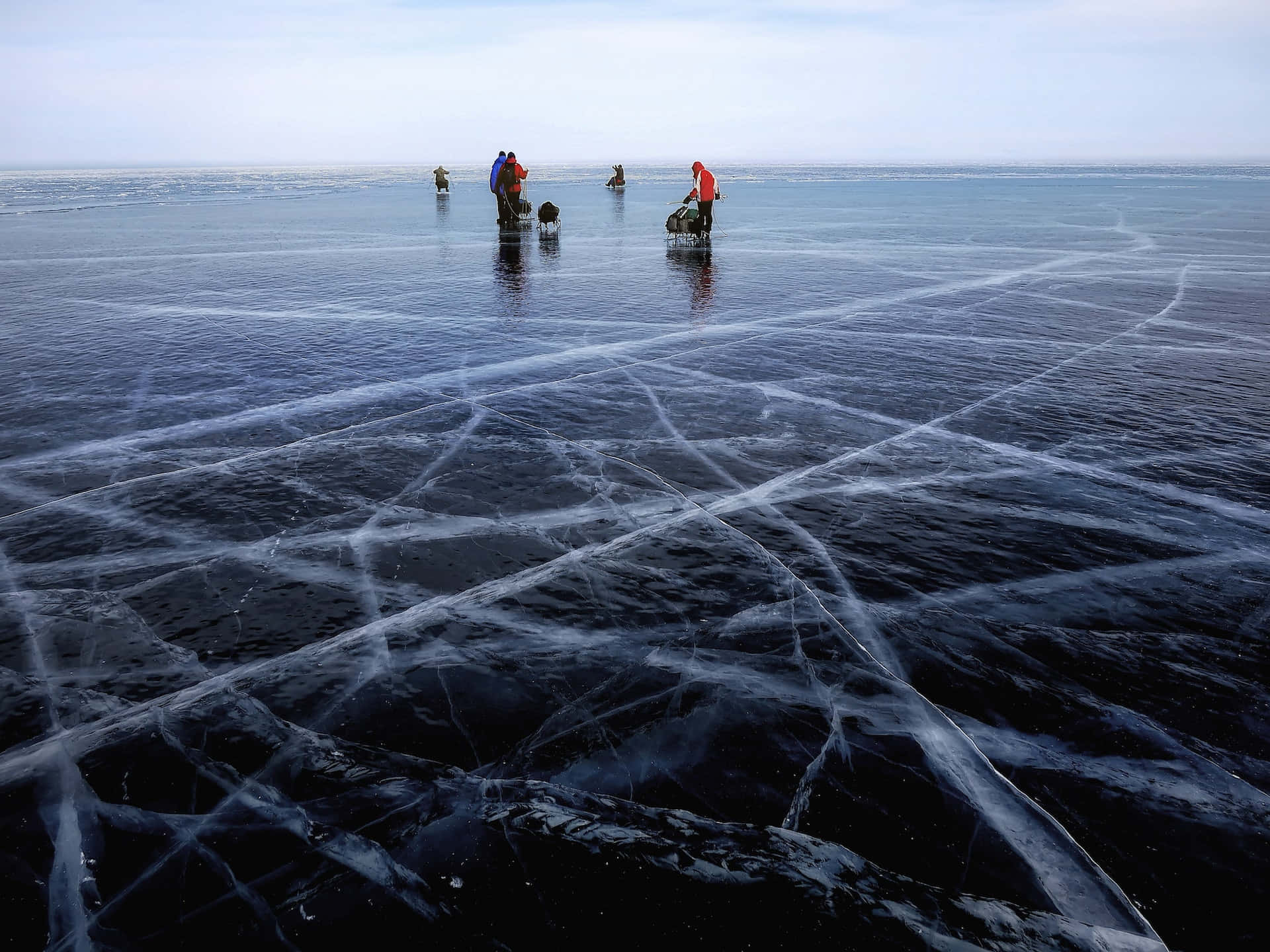 Ice Fishing 桌布 壁紙 — 人們站在廣闊的冰凍湖面上，複雜的冰裂紋反射著蒼白的天空。