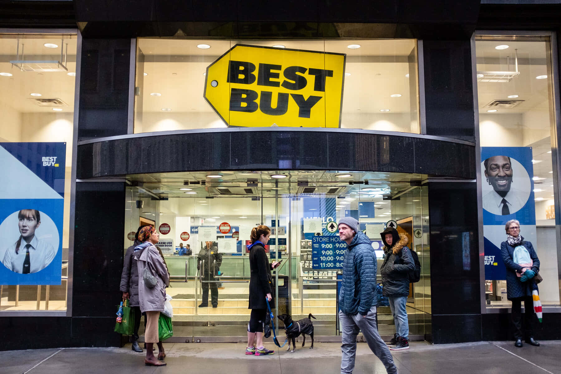 Best Buy 桌布 壁紙 — 人們聚集在Best Buy商店入口外，入口有明亮的黃色標誌和玻璃門，反射著城市燈光。