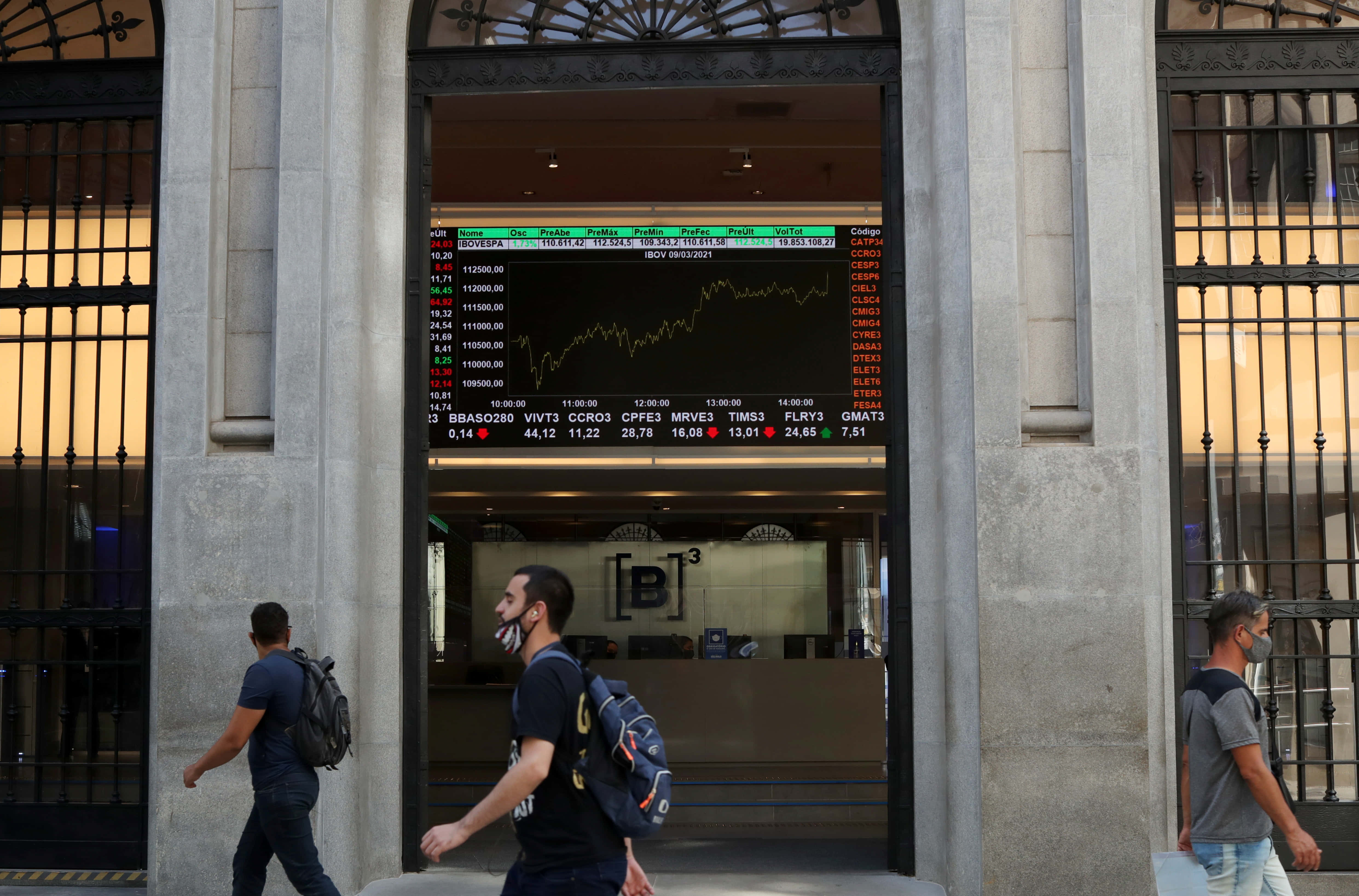 Stock 桌布 壁紙 — 人們走過一個大型石造建築入口，顯示一個發光的股票市場行情板，上面有波動的金融數據。
