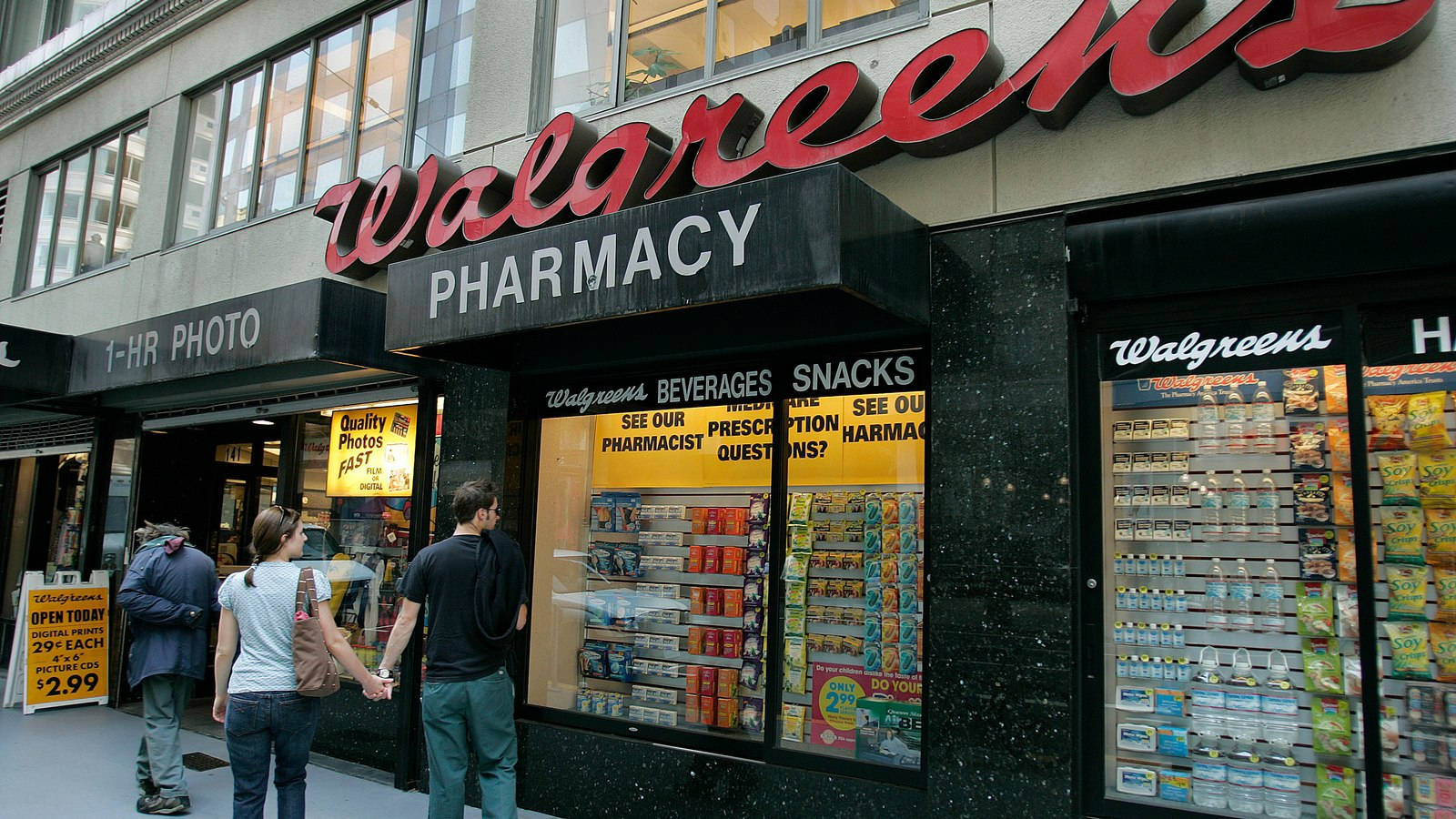 Business 桌布 壁紙 — 人們走過一間Walgreens藥房，店面有鮮紅色標誌和裝滿零食飲品的玻璃陳列櫃。
