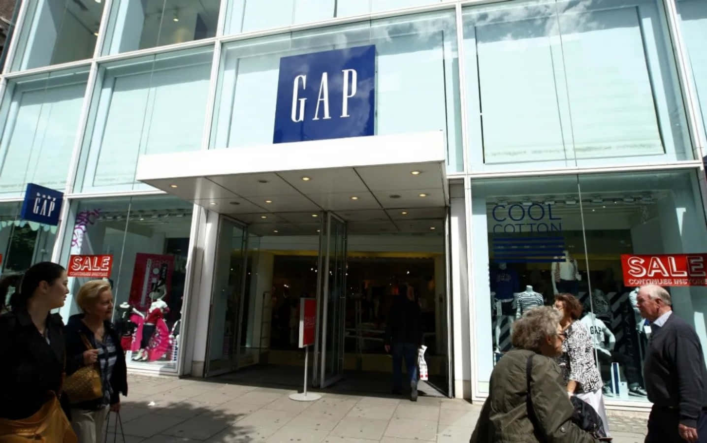 Gap 桌布 壁紙 — 人們走過一間現代玻璃幕牆的GAP商店，可見減價標誌和顧客進入。