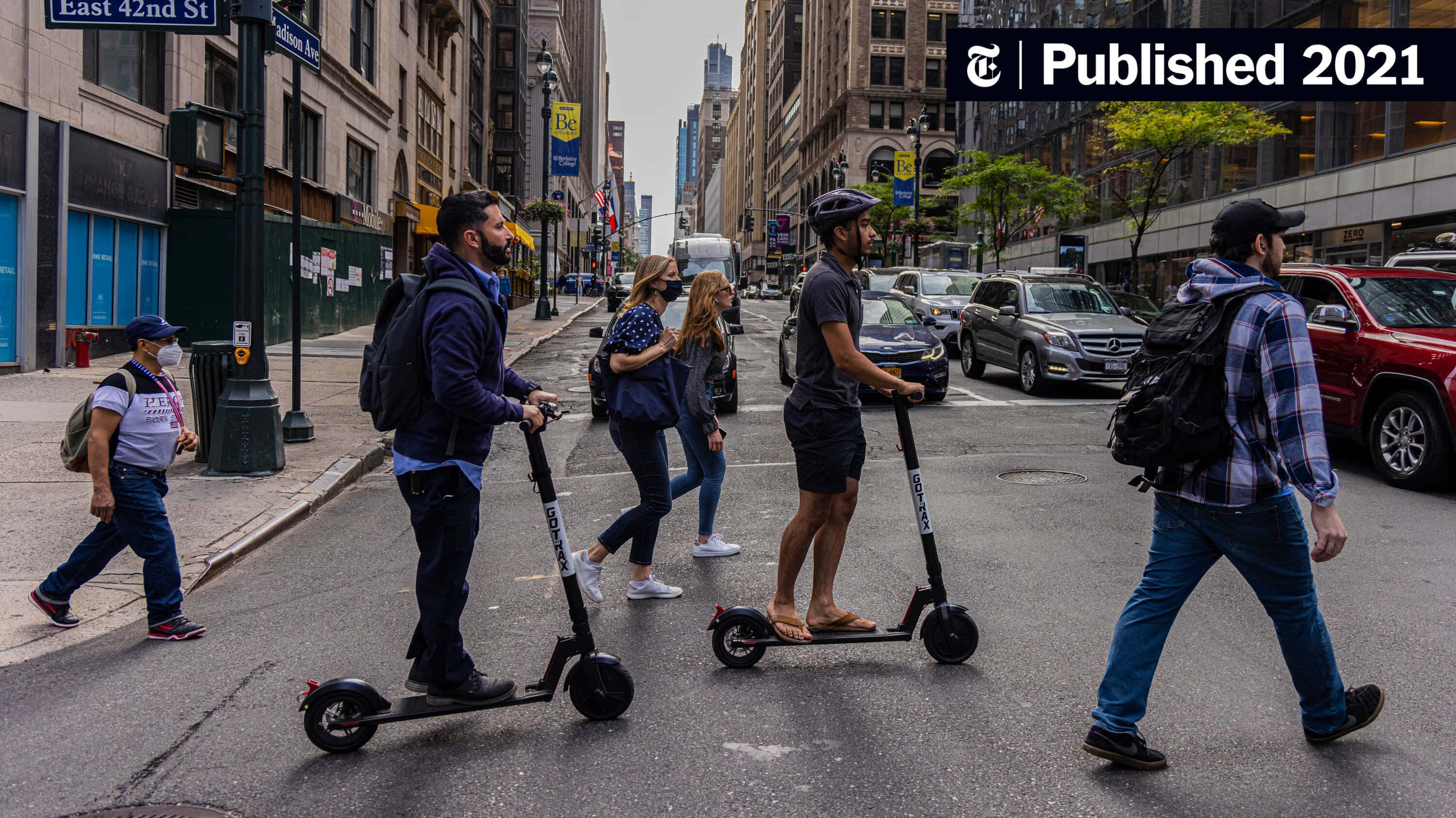 scooter 桌布 壁紙 — 人們騎著電動滑板車和步行穿過一條繁忙的城市街道，兩旁是高樓大廈和汽車。