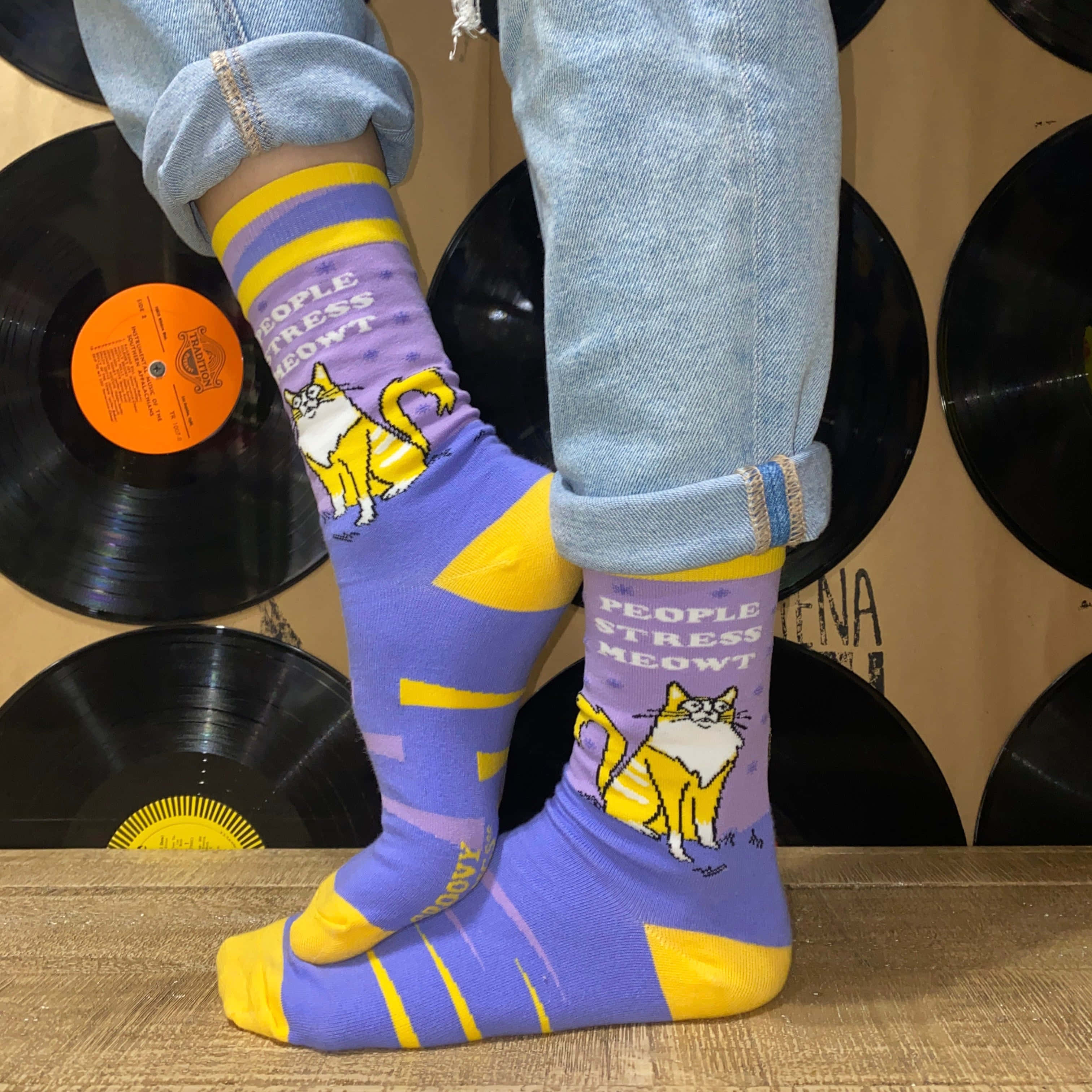 Purple Socks 桌布 壁紙 — 人物穿著紫色和黃色襪子，上面有卡通貓和文字，站在木地板上，背景是黑膠唱片牆。