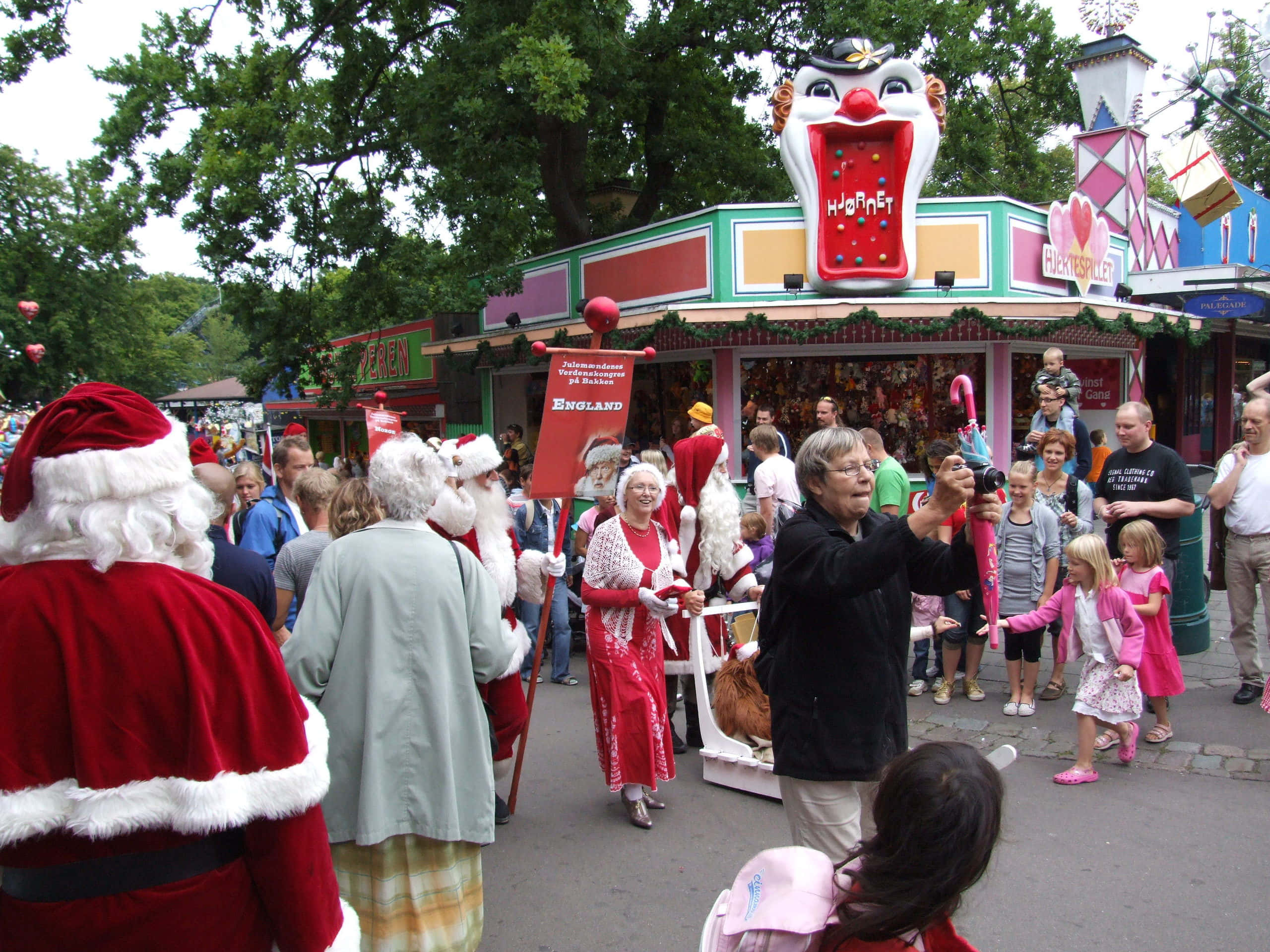 Parade 桌布 壁紙 — 人群包括聖誕老人和兒童聚集在一個色彩繽紛的遊樂園攤位周圍，攤位有一個巨大的小丑頭。