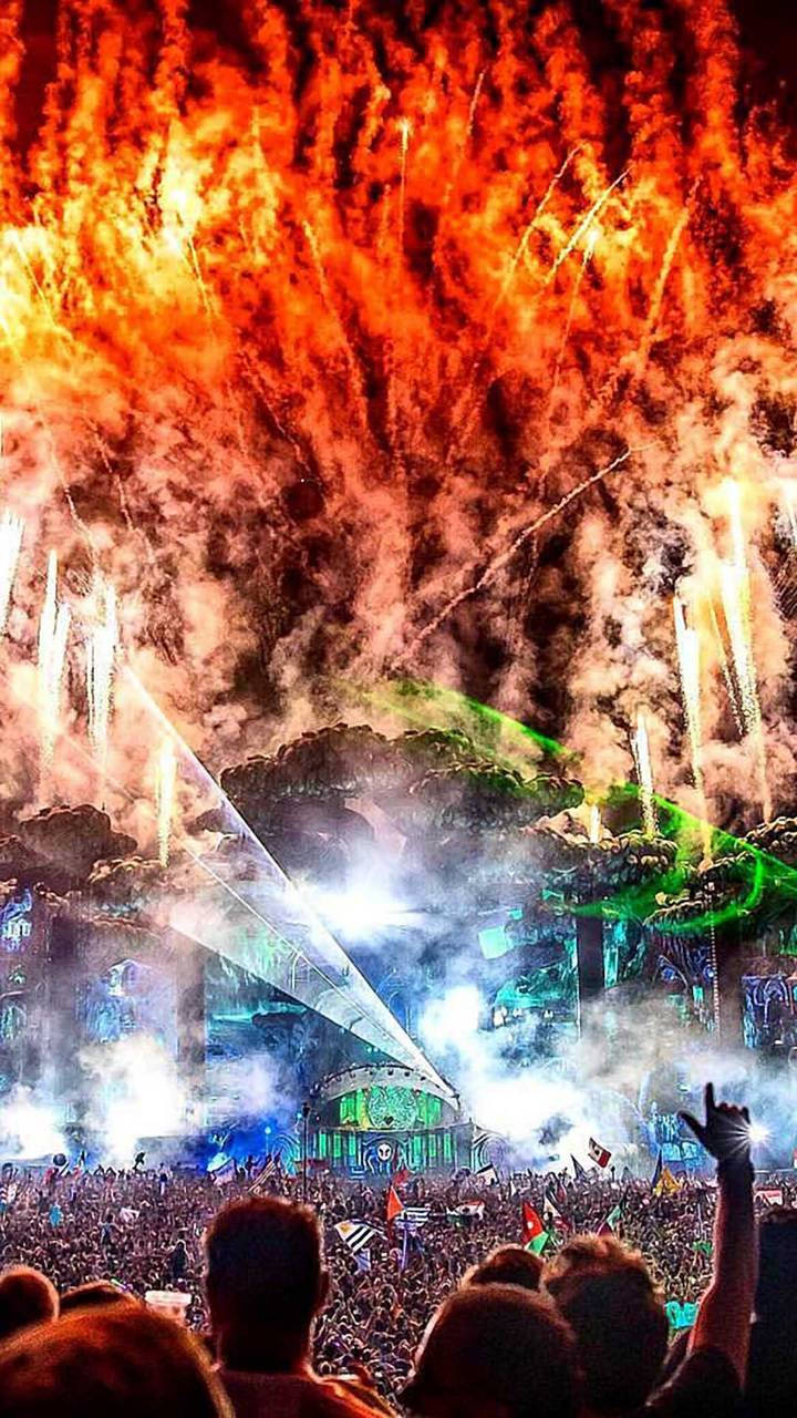 Tomorrowland 桌布 壁紙 — 人群向上湧動，熾熱的橙色煙火在發光的綠色舞台上空爆炸，這是一場大型夜間節日。