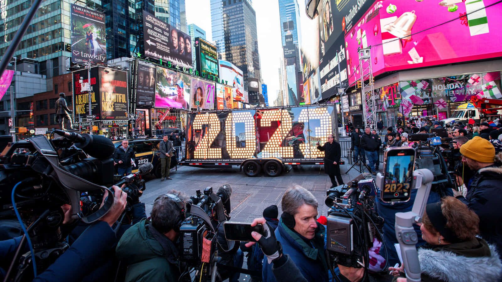 New York 桌布 壁紙 — 人群和相機聚集在時代廣場一輛閃閃發光的2023年遊行花車周圍，摩天大樓和廣告牌照亮了繁華的城市景象。