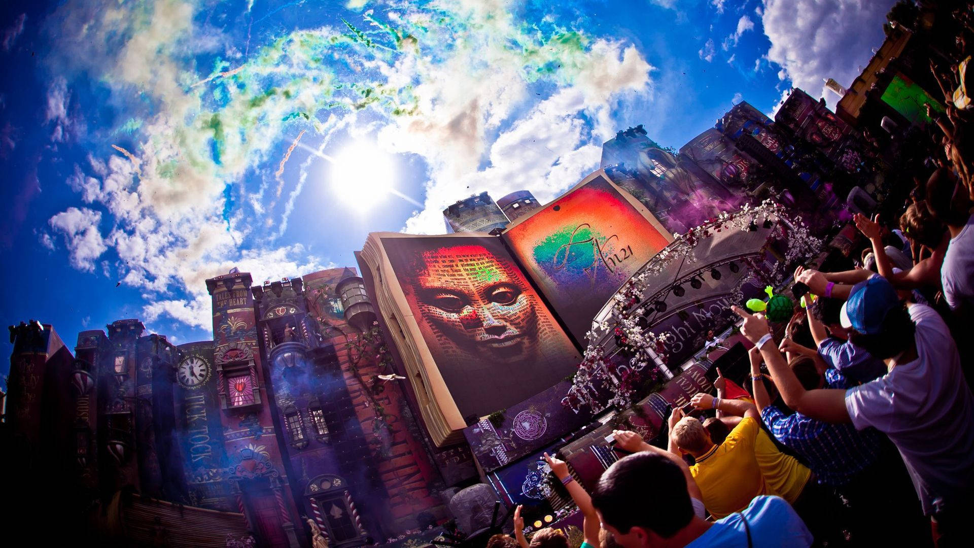 Tomorrowland 桌布 壁紙 — 人群在充滿活力的天空下歡呼，巨大的書本舞台配上色彩繽紛的臉部投影，主導了節日場景。