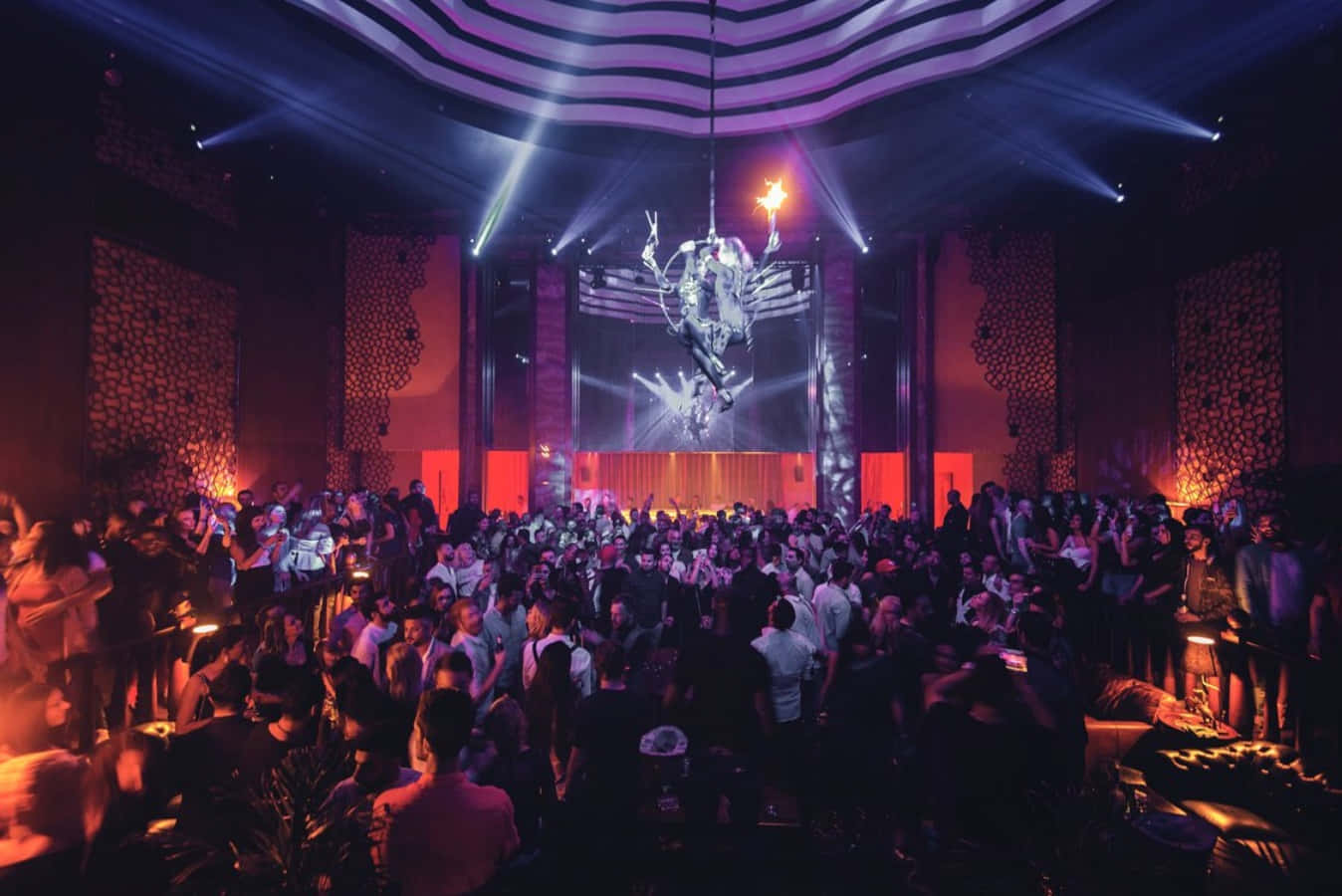 club 桌布 壁紙 — 人群在充滿活力的紫色和橙色燈光下，在擠滿人的夜總會中跳舞，現場有發光的吊燈和舞台。