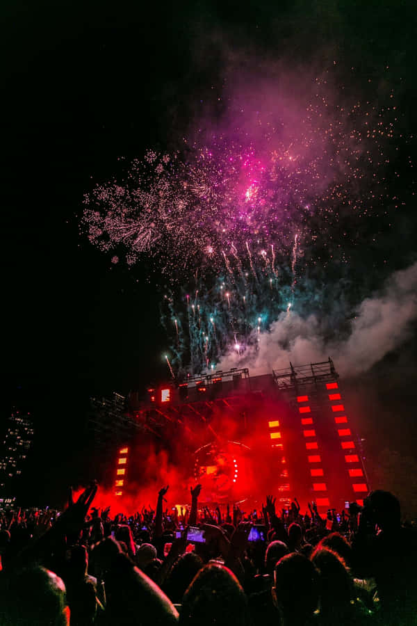 concert 桌布 壁紙 — 人群在夜晚紅色燈光的演唱會舞台上，於耀眼的粉紅色和藍綠色煙火爆炸中歡呼。