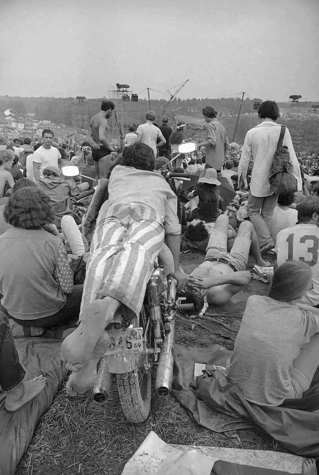 woodstock 桌布 壁紙 — 人群在戶外音樂節的草地上放鬆，背景有摩托車和舞台。
