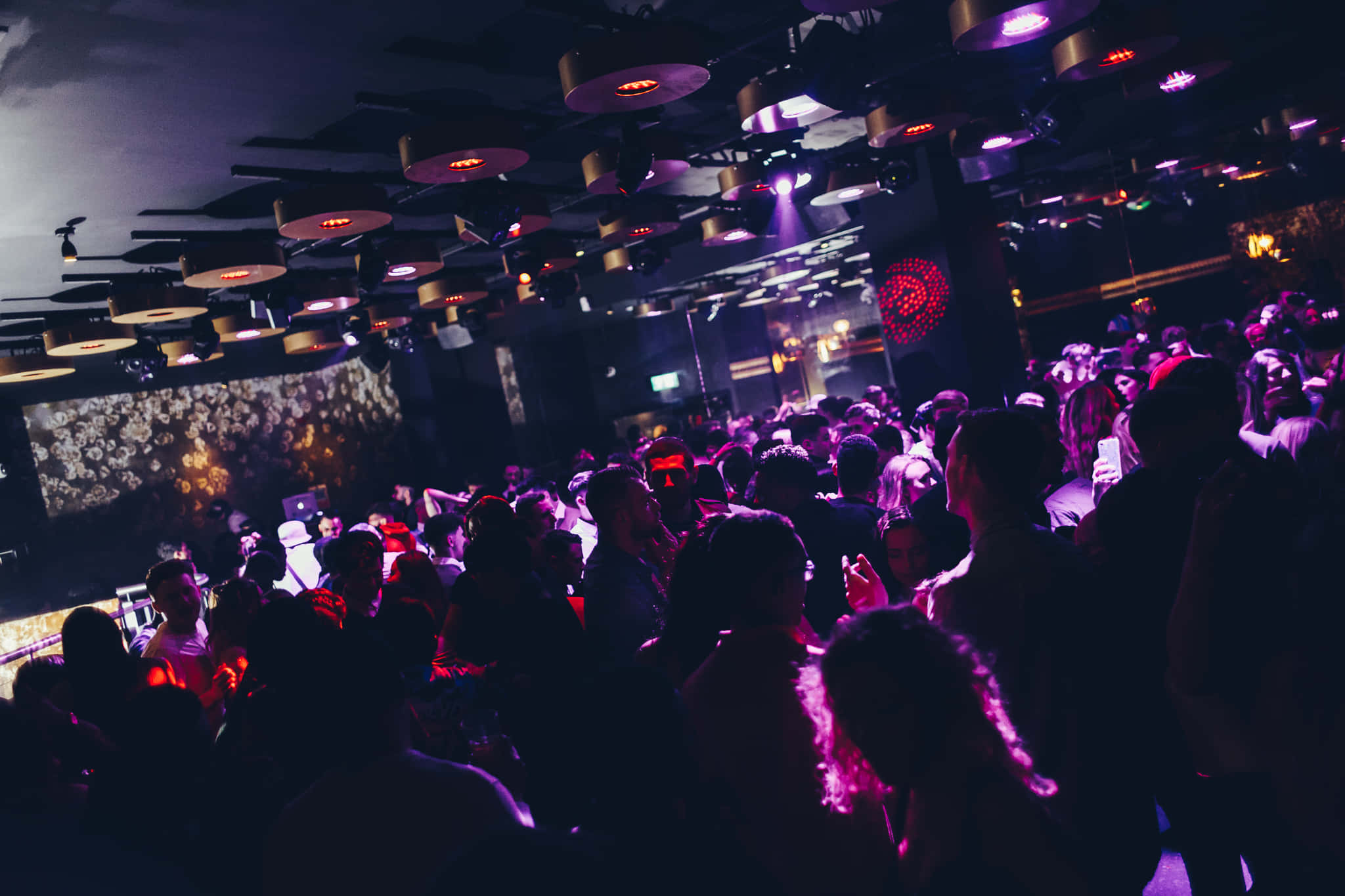 club 桌布 壁紙 — 人群在昏暗的夜店中，於脈動的紫色燈光、天花板風扇和發光舞台裝飾下跳舞。