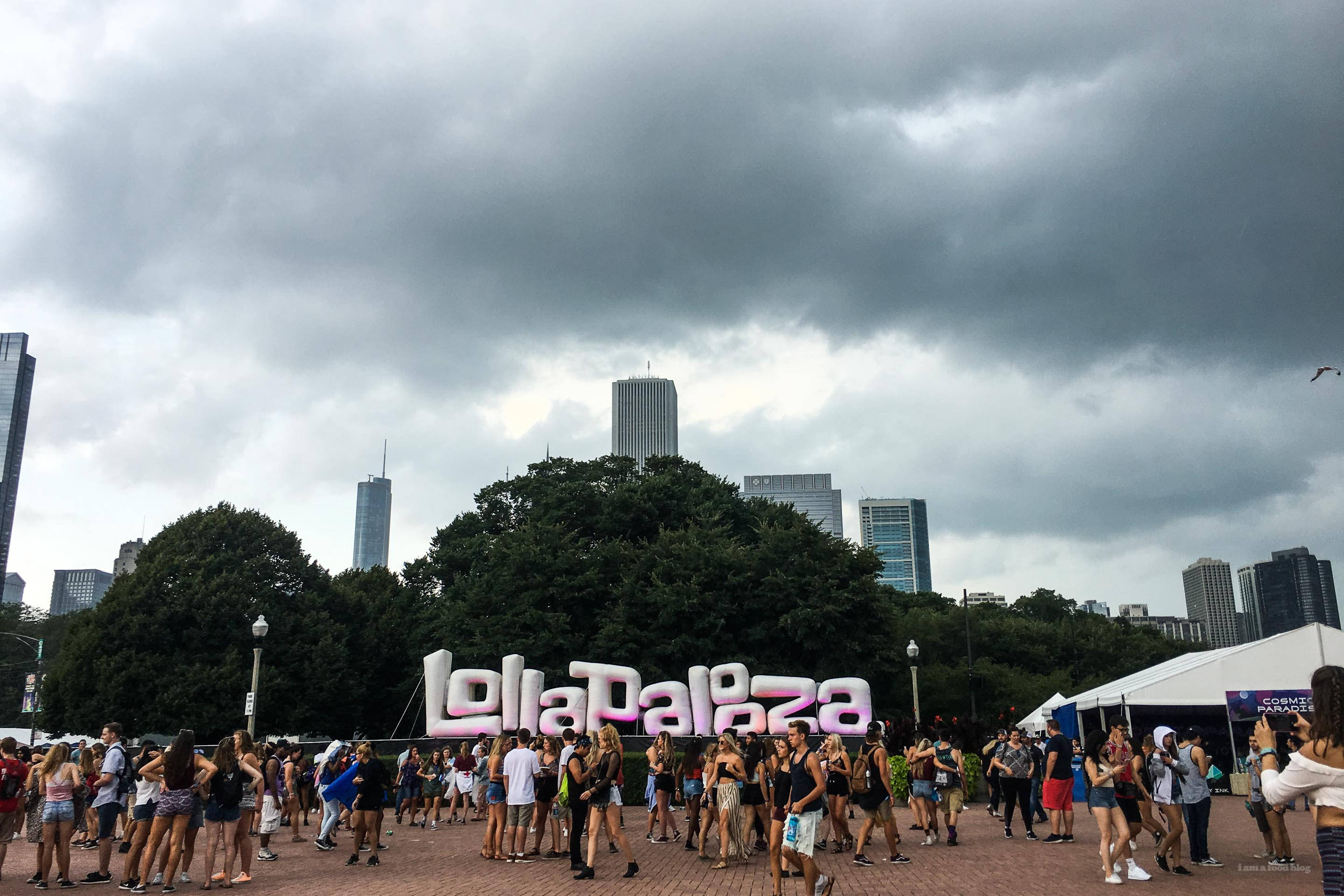 Lollapalooza 桌布 壁紙 — 人群在暴風雨的天空下聚集在一個巨大的Lollapalooza標誌周圍，背景是城市摩天大樓和樹木。