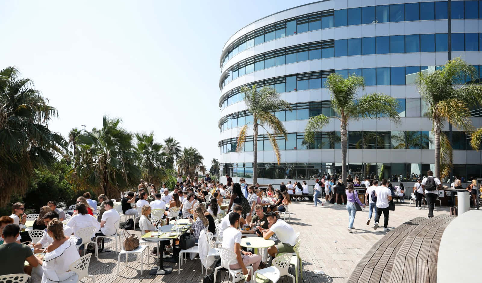 Nice 桌布 壁紙 — 人群在現代玻璃建築和棕櫚樹旁的戶外咖啡館露台上享受陽光明媚的一天。