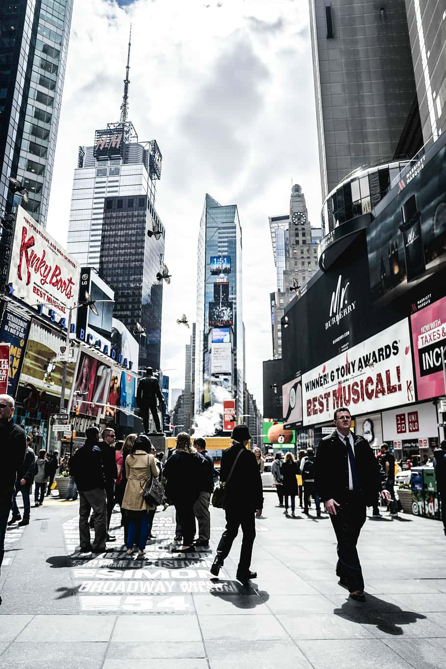 new york city iPhone x 桌布 壁紙 — 人群在紐約市一條繁華的街道上穿梭，街道兩旁高聳的摩天大樓和充滿活力的百老匯劇院廣告牌在陰天之下。