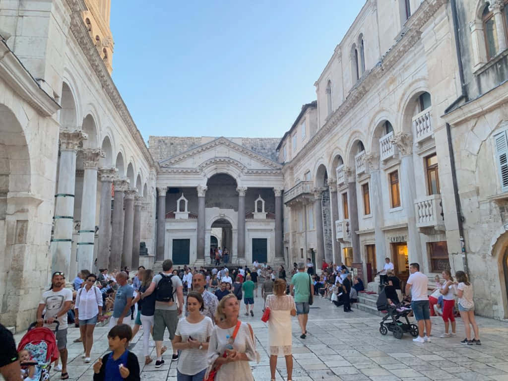 Croatia 桌布 壁紙 — 人群在陽光普照的歷史廣場漫步，兩旁是帶有拱形柱廊和陽台的宏偉石建築。