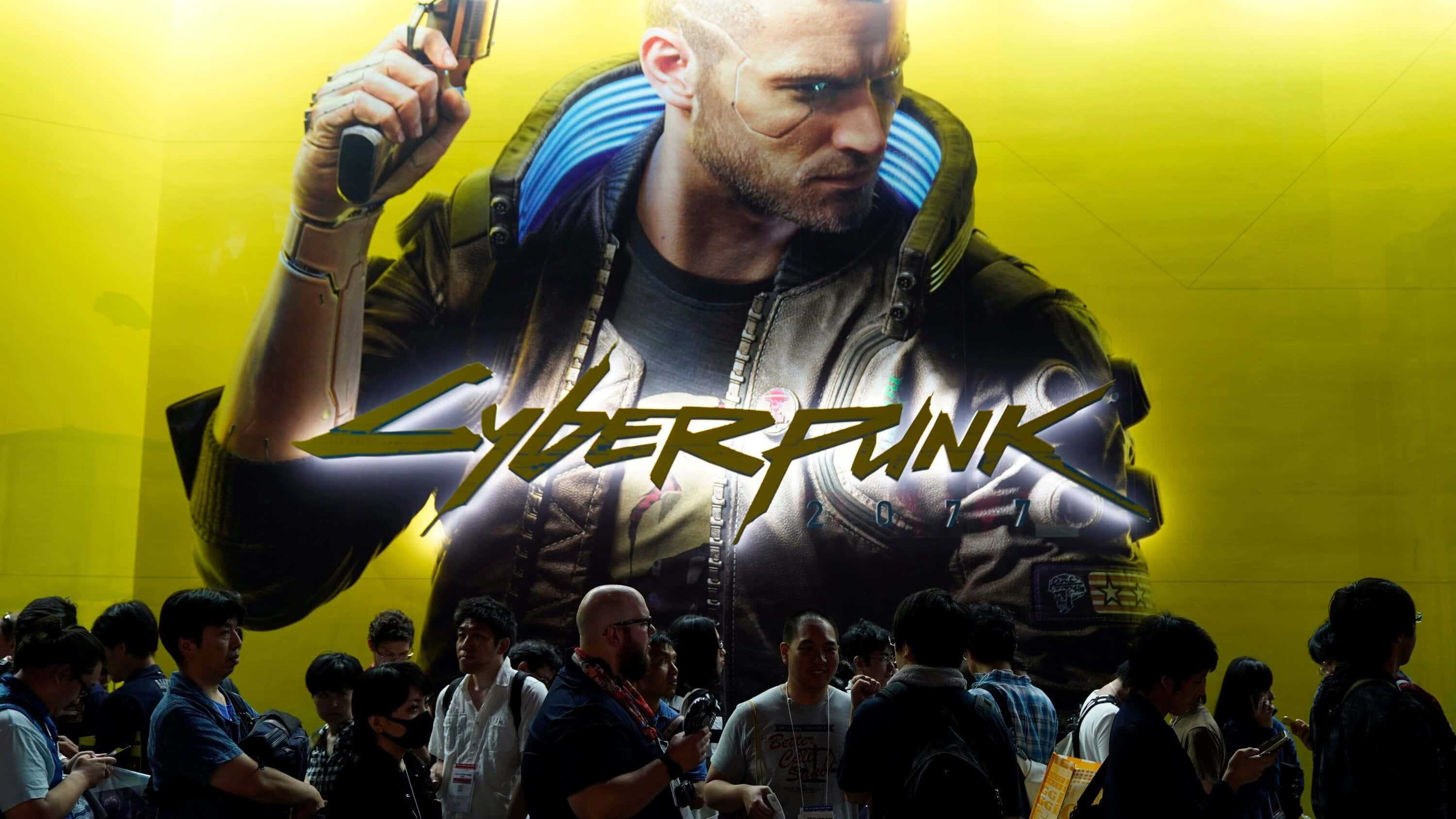 賽博龐克 桌布 壁紙 — 人群聚集在一個巨大的發光黃色 Cyberpunk 2077 海報前，海報上有一個機械人拿著槍。