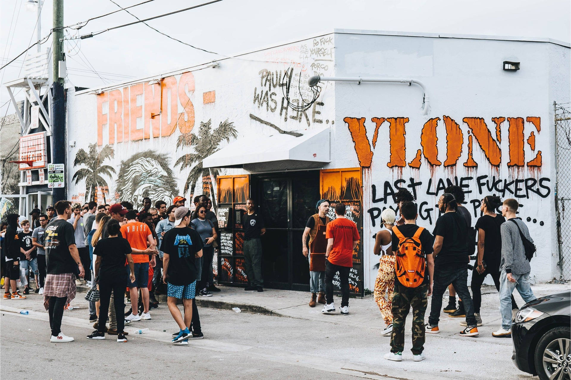Vlone 桌布 壁紙 — 人群聚集在一座被塗鴉覆蓋的建築物外，建築物上有大膽的橙色和黑色街頭藝術，天空多雲。