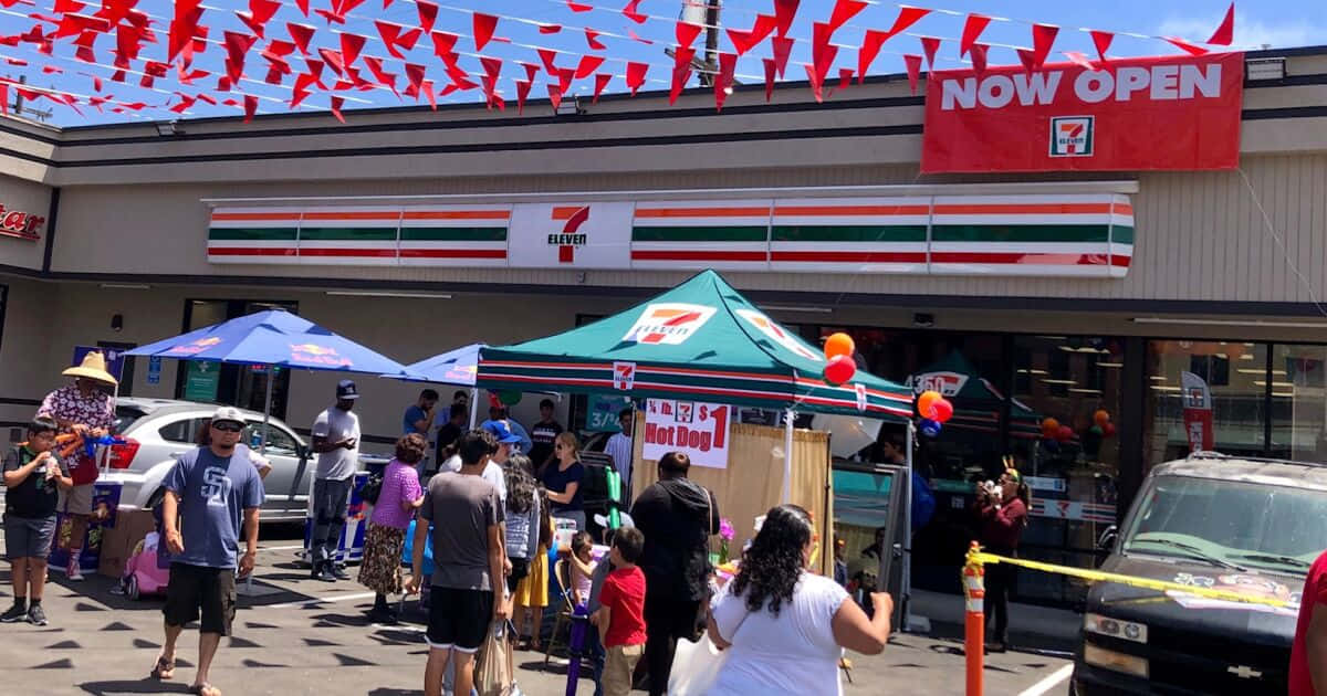 7-ELEVEN 桌布 壁紙 — 人群聚集在一間新開幕的7 eleven商店外，上方有紅色三角旗幟和明亮的藍天。