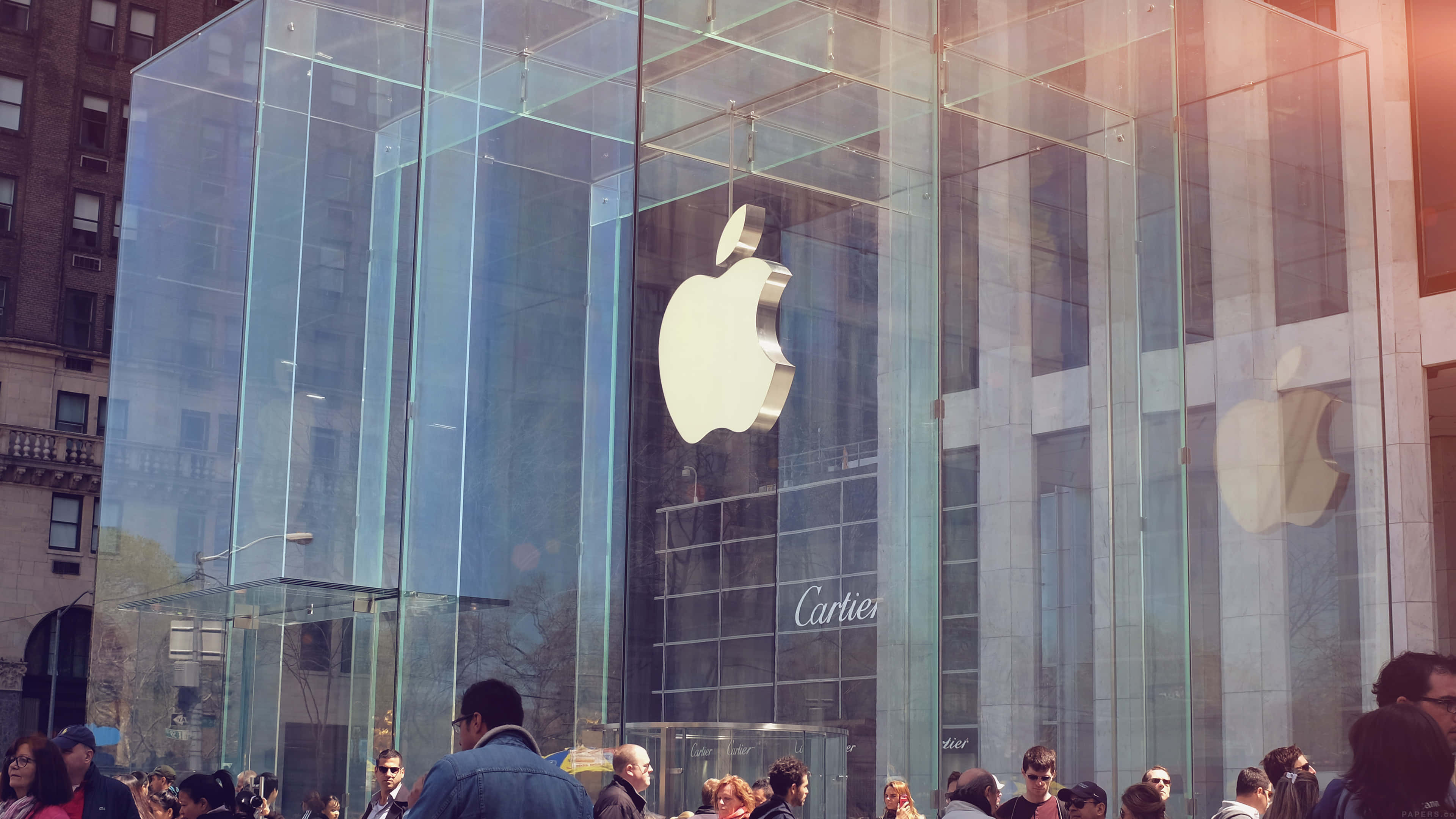 桌布 壁紙 — 人群聚集在時尚的玻璃Apple Store外，一個大型銀色標誌反射著陽光普照的城市景觀。