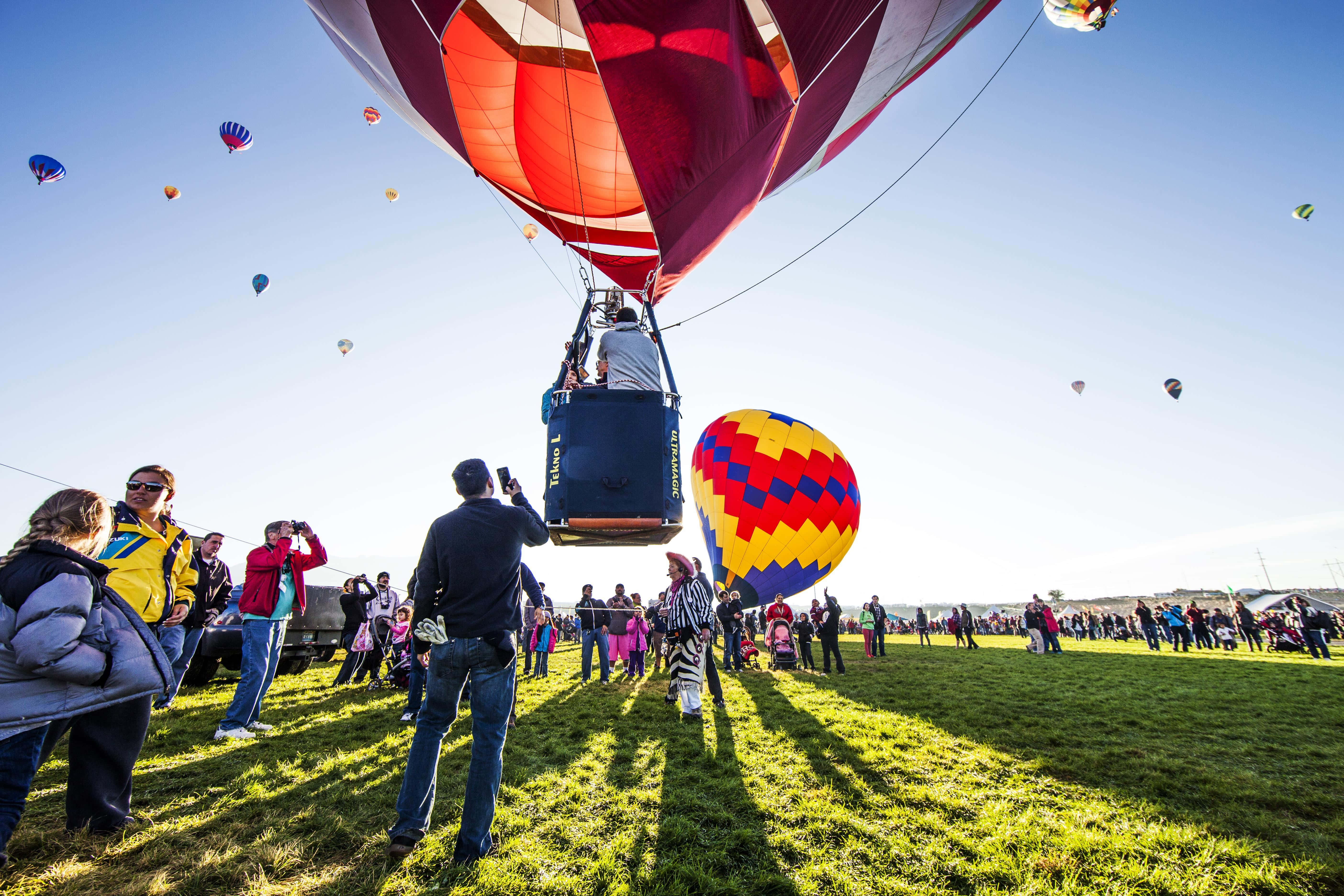 Hot Air Balloon 桌布 壁紙 — 人群聚集在陽光照耀的草地上，觀賞色彩繽紛的熱氣球升上清澈的藍天。