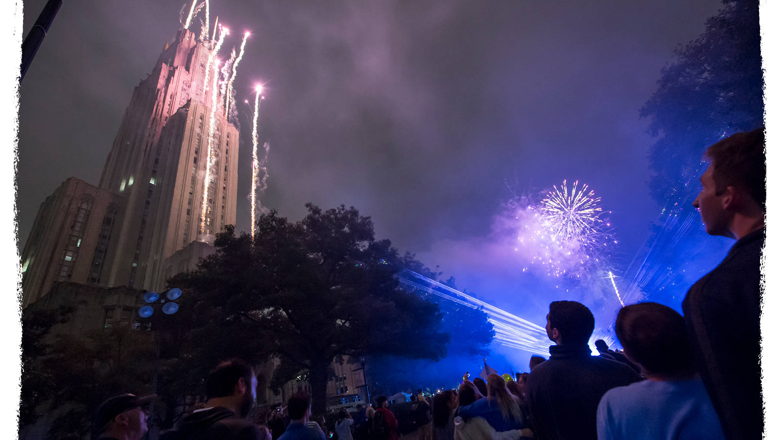 Fireworks 桌布 壁紙 — 人群觀看令人驚嘆的紫色煙火在夜空下高聳的照明建築上方爆炸。