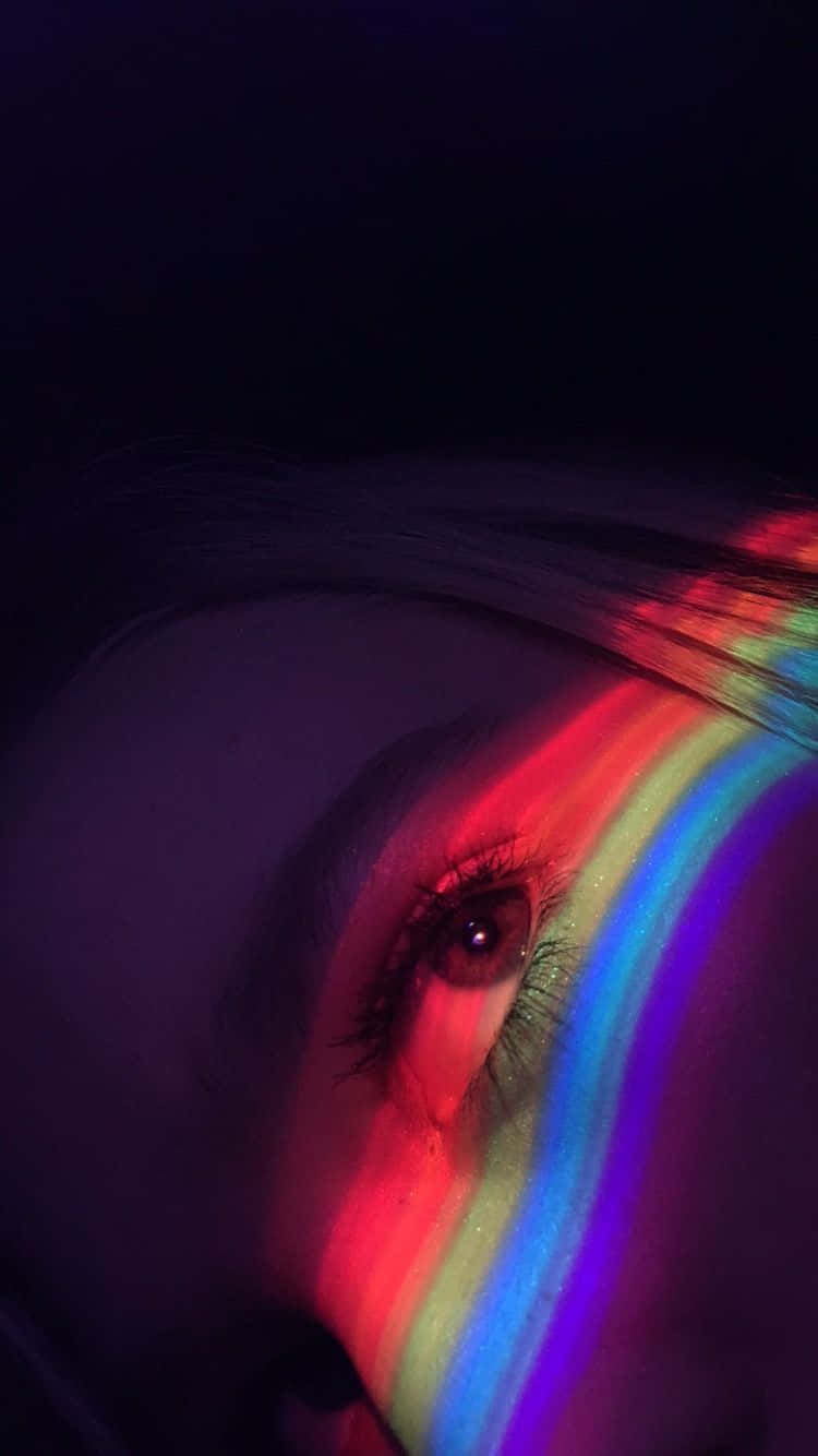 彩虹美學 桌布 壁紙 — 人類眼睛的特寫，被鮮豔的彩虹光照亮，在黑暗背景上於臉頰和頭髮上投射出色彩繽紛的條紋。