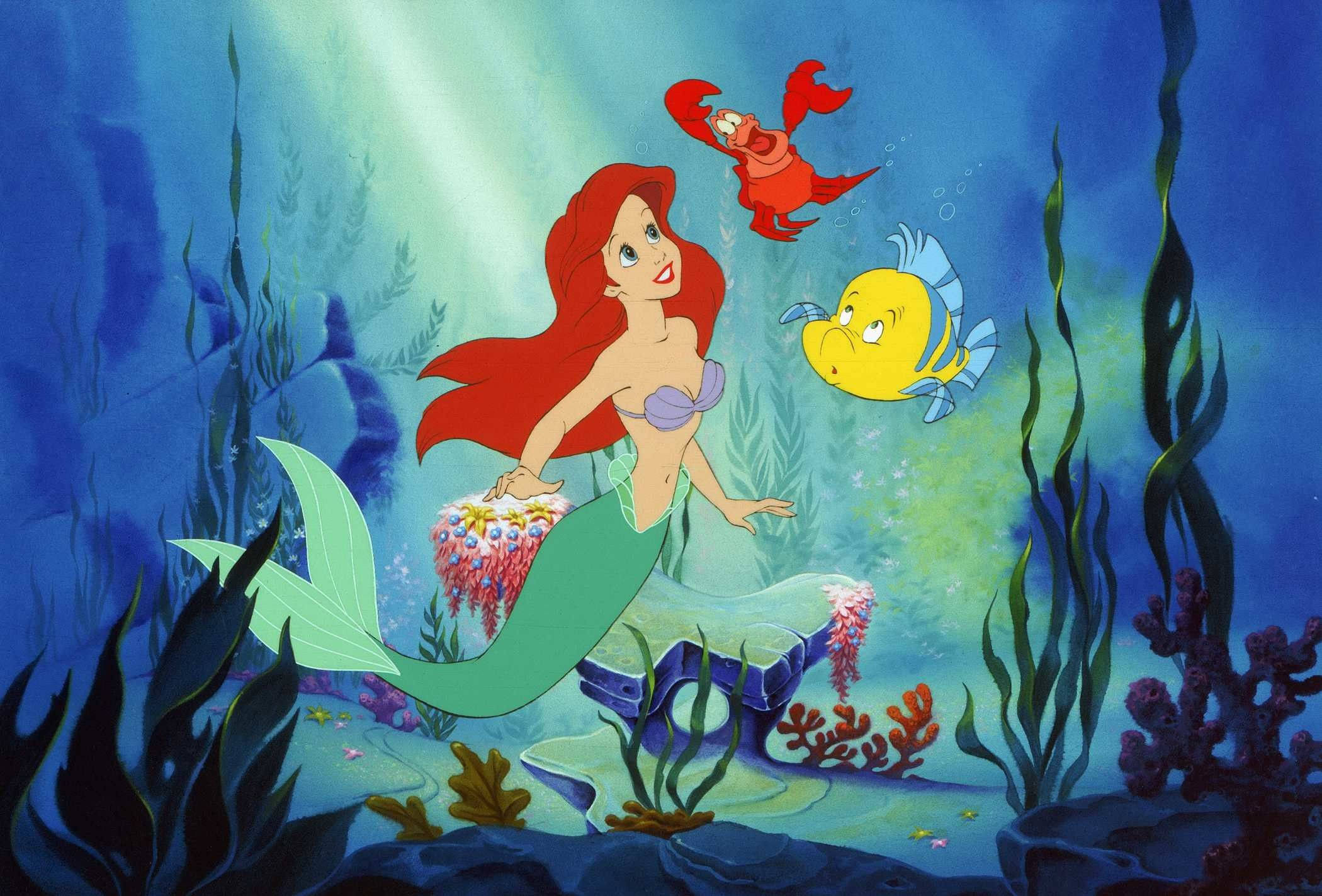 The Little Mermaid 桌布 壁紙 — 人魚愛麗兒（Ariel）擁有一頭飄逸的紅髮，坐在珊瑚礁上微笑，她的螃蟹朋友和魚兒同伴在陽光普照的海水中游近。