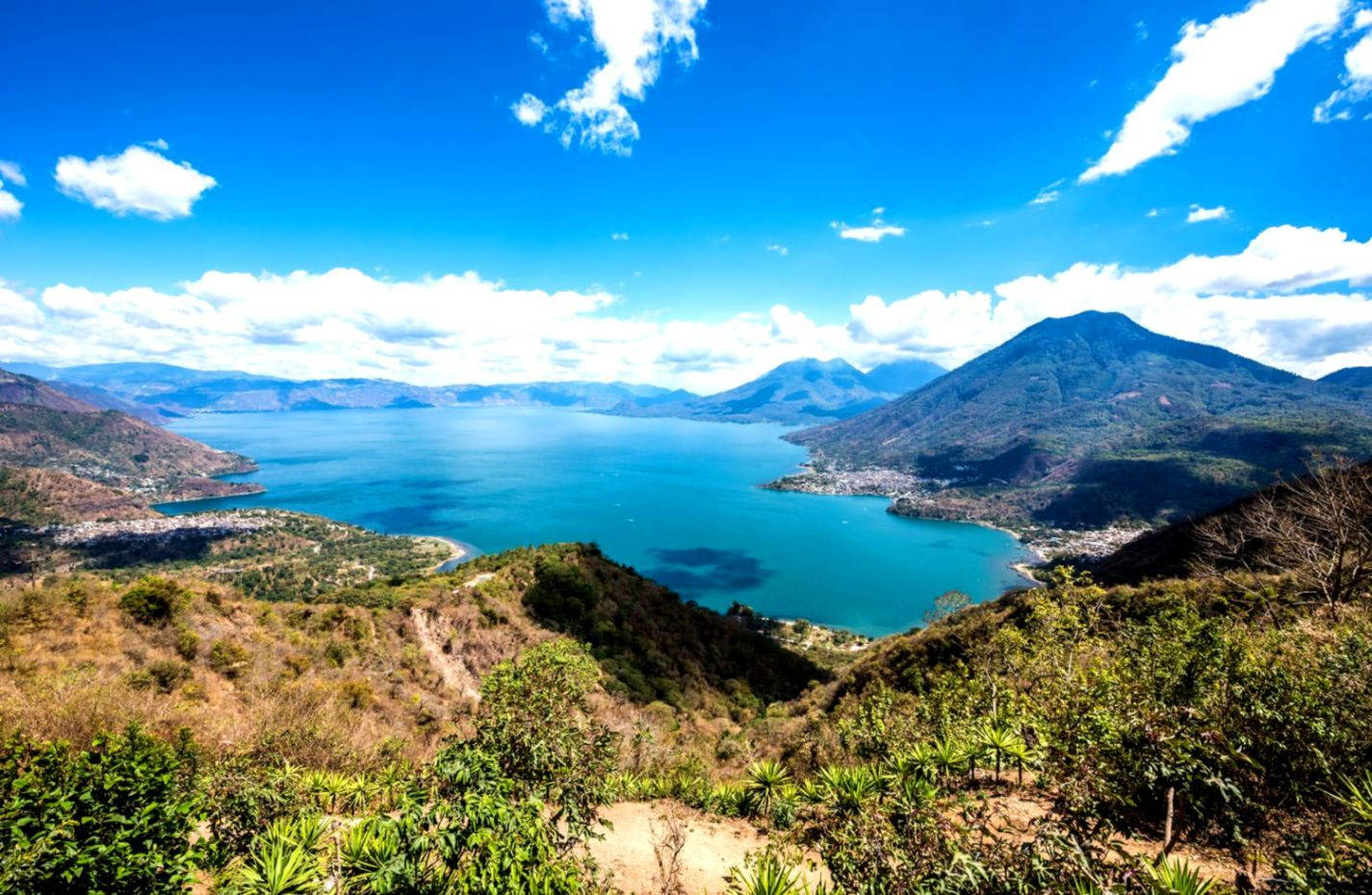 Guatemala 桌布 壁紙 — 令人嘆為觀止的全景視野，一個碧綠色的湖泊 nestled 在鬱鬱蔥蔥的綠色山脈之間，在明亮的藍天和 fluffy 白雲下。