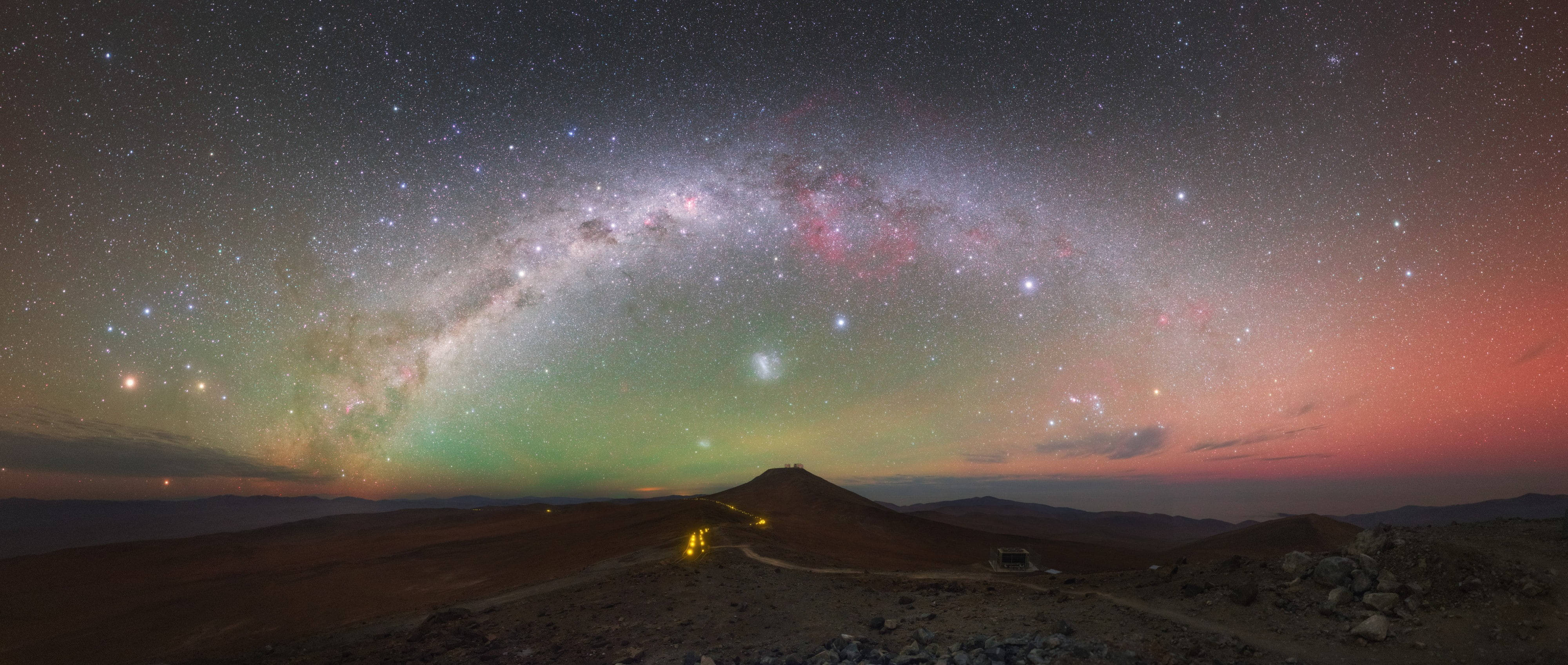 Chile 桌布 壁紙 — 令人嘆為觀止的全景視野，銀河在暮色中拱起於沙漠景觀之上，沿著蜿蜒的道路有充滿活力的星光和遠處的燈光。