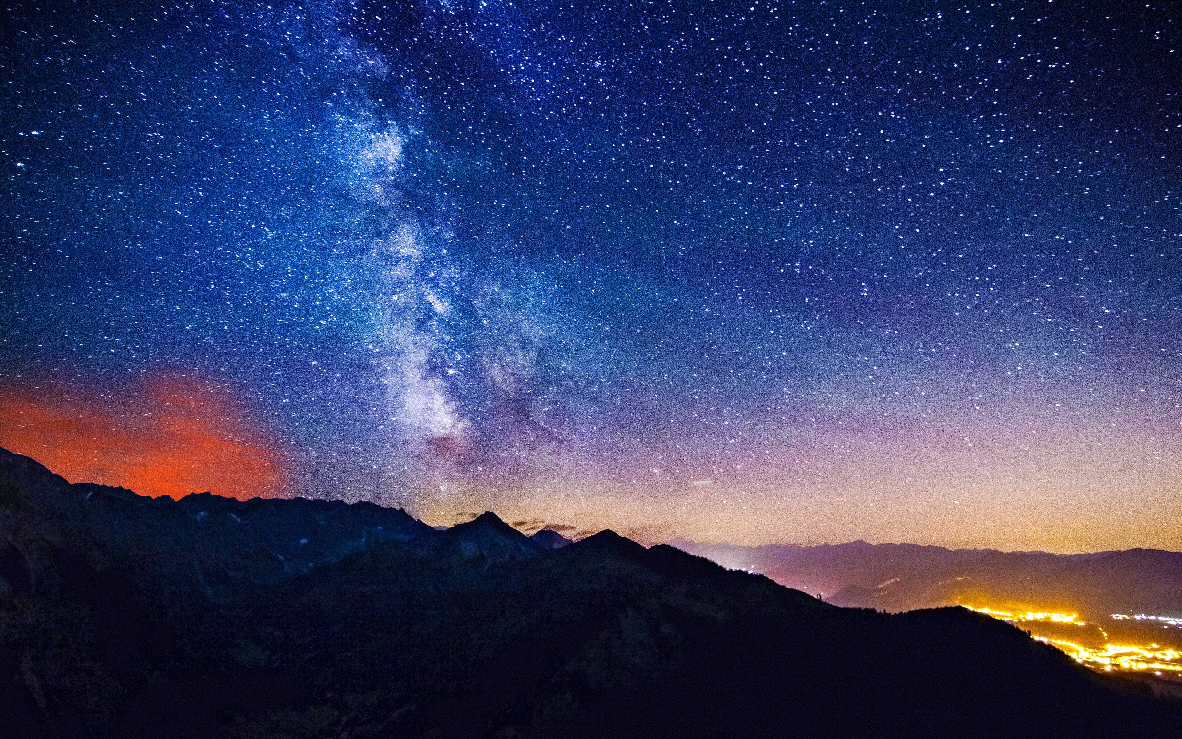 宇宙美學 桌布 壁紙 — 令人嘆為觀止的夜空，佈滿星星，銀河在黑暗的山巒剪影上拱起，遠處的城市燈光在下方閃爍。