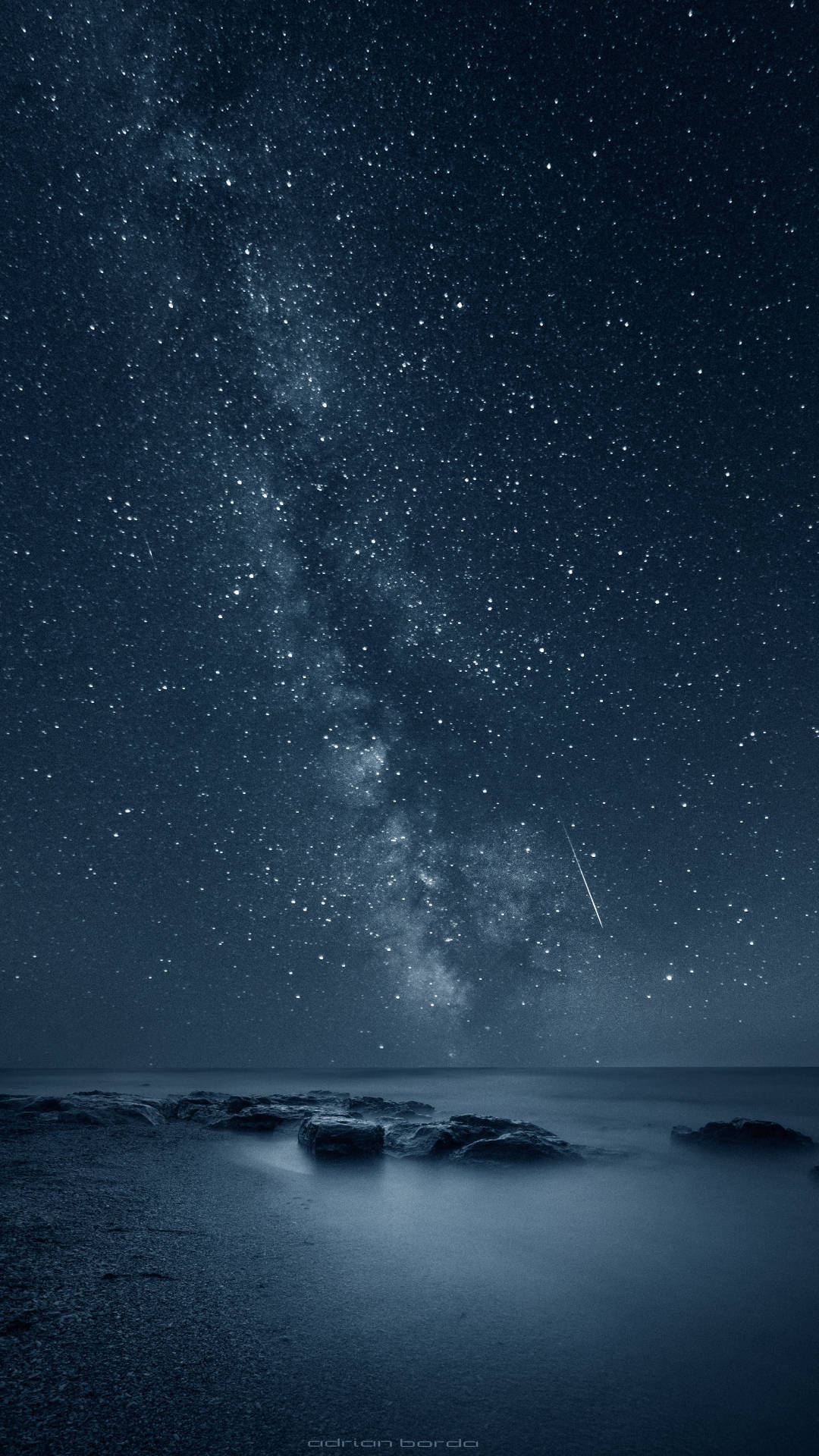 iPhone 桌布 壁紙 — 令人嘆為觀止的夜空，佈滿繁星和銀河，拱形橫跨在平靜的海洋上，前景有深色岩石。