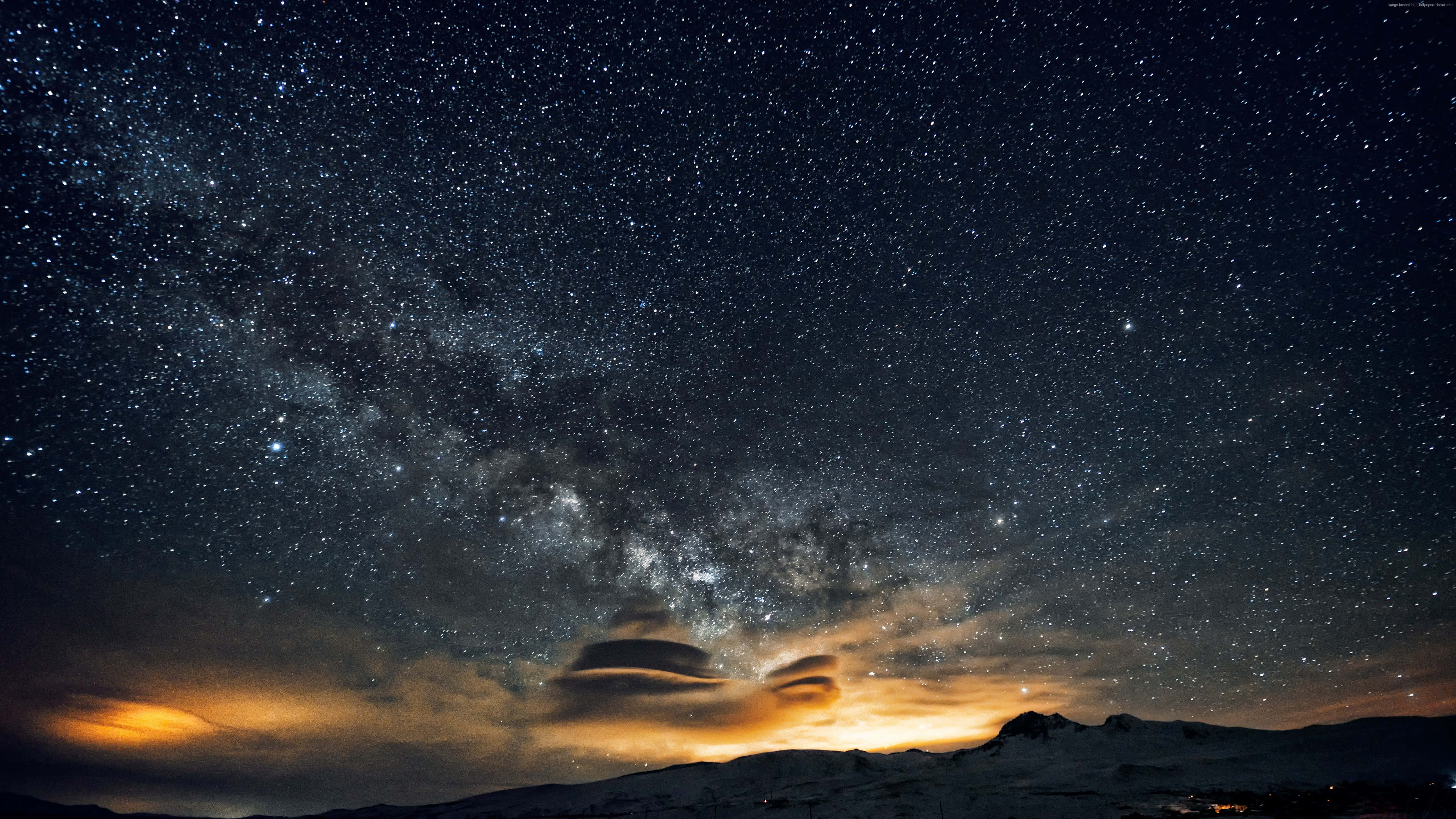 Trippy Galaxy 桌布 壁紙 — 令人嘆為觀止的夜空，充滿無數星星和銀河，拱形跨越白雪覆蓋的山脈，下方有發光的橙色雲彩。