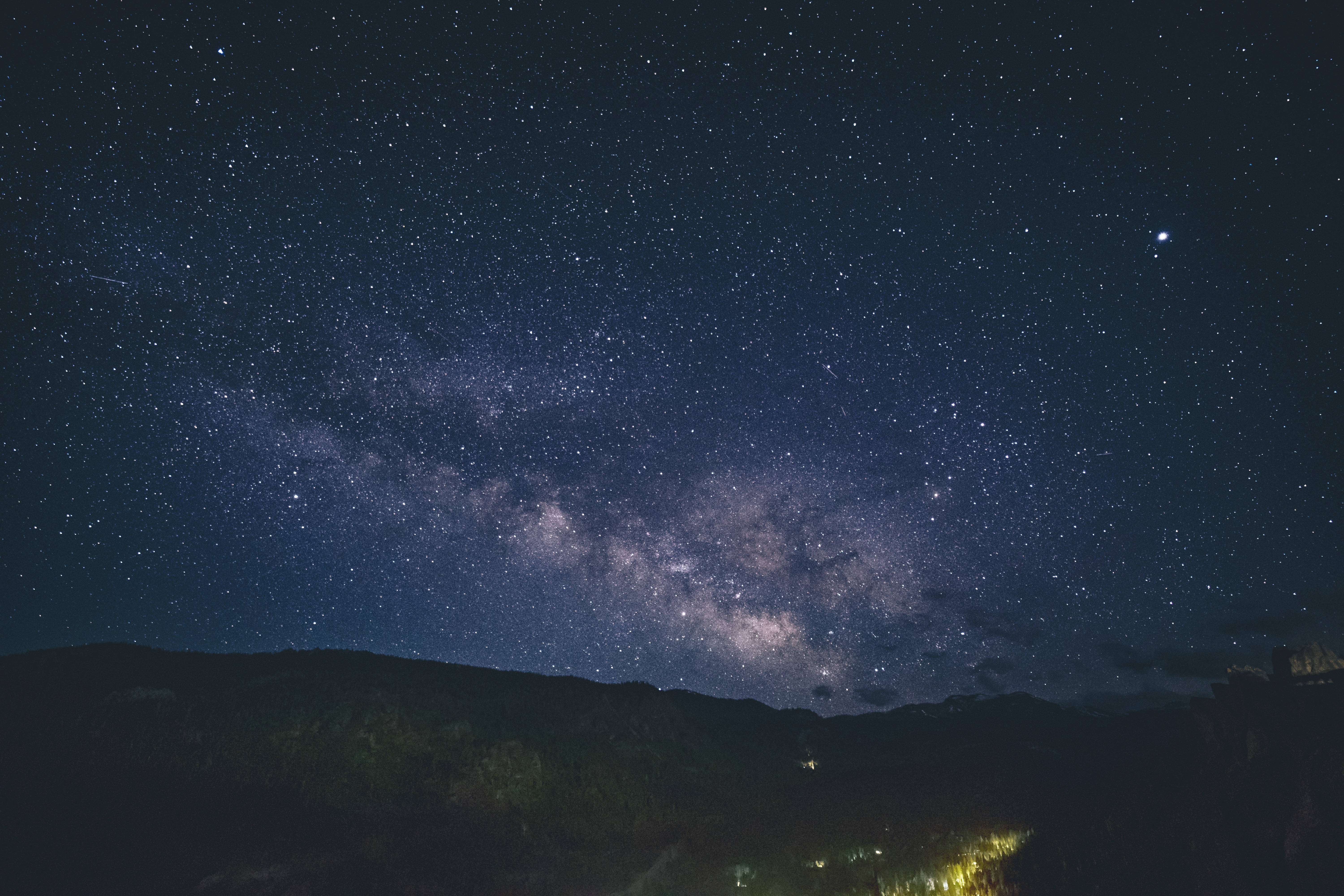 藍色銀河 桌布 壁紙 — 令人嘆為觀止的夜空，充滿無數星星，發光的銀河在黑暗的山脈上方拱起，下方有遙遠的村莊燈光。