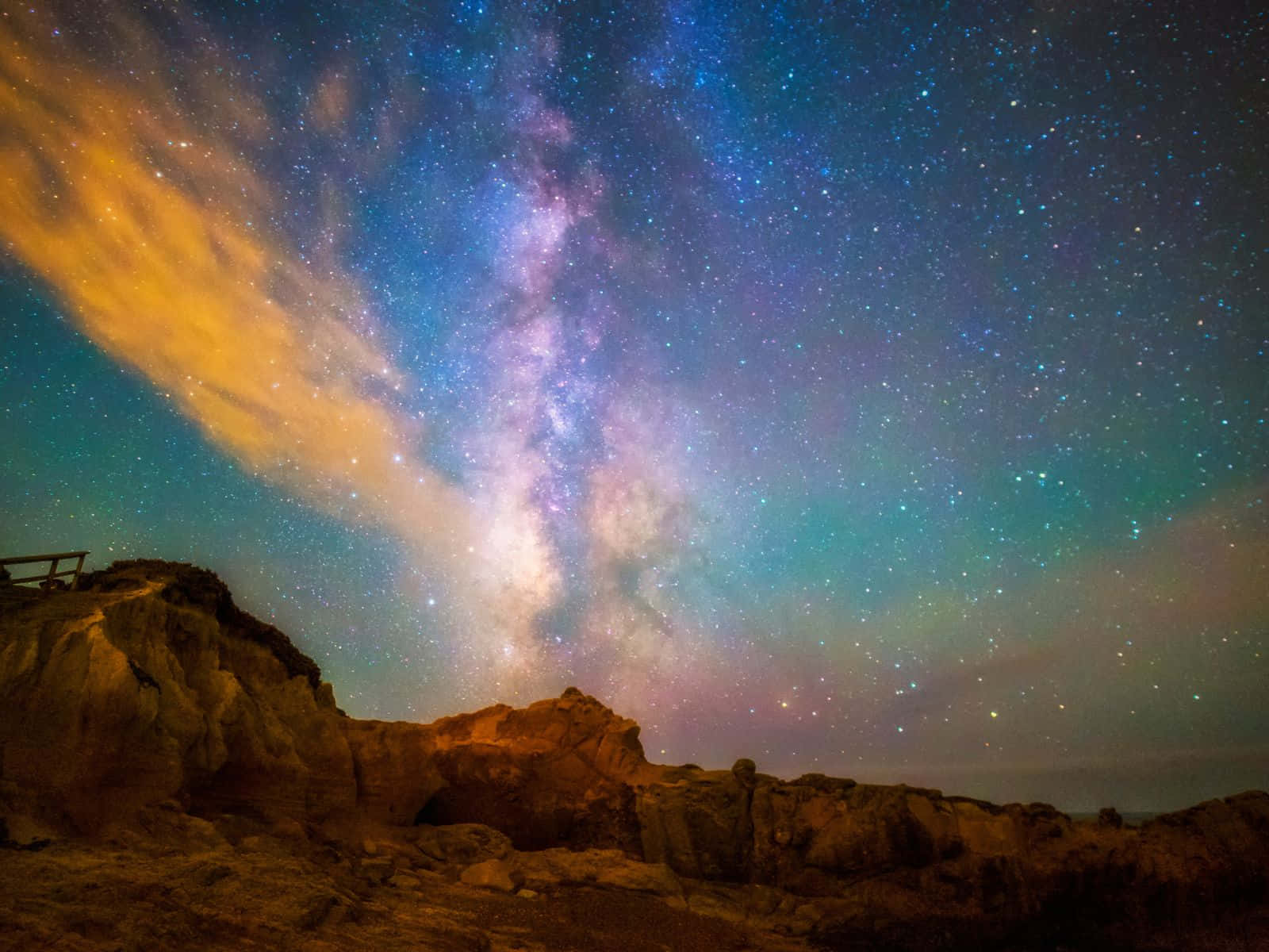 1600x1200 桌布 壁紙 — 令人嘆為觀止的夜空，滿佈繁星和銀河，在發光的橙色雲朵下拱起於崎嶇的岩石地形之上。