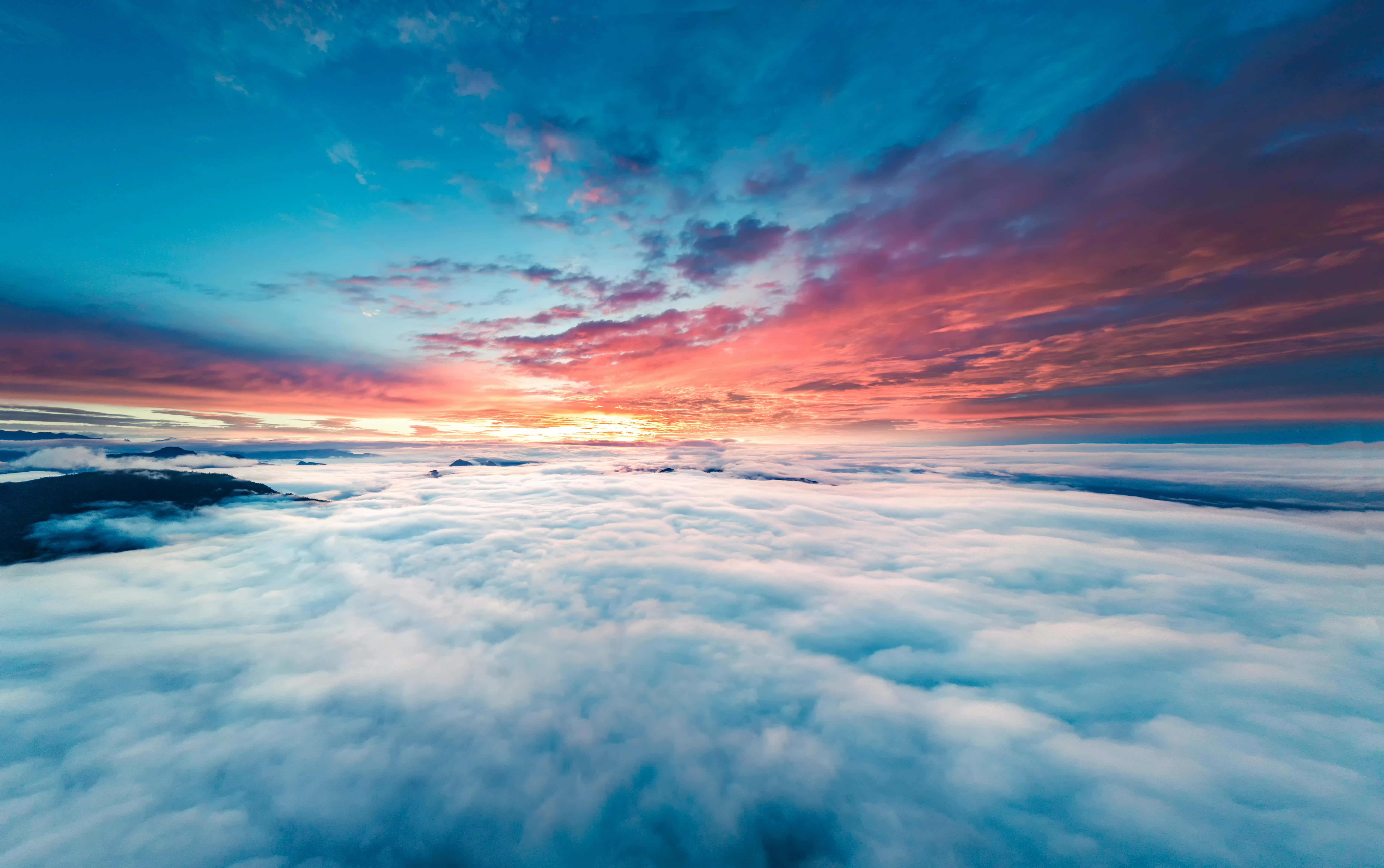 Clouds Aesthetics 桌布 壁紙 — 令人嘆為觀止的日出，在蓬鬆白雲的海洋之上，以火熱的橙色和深藍色描繪天空，遠方有山巒的剪影。