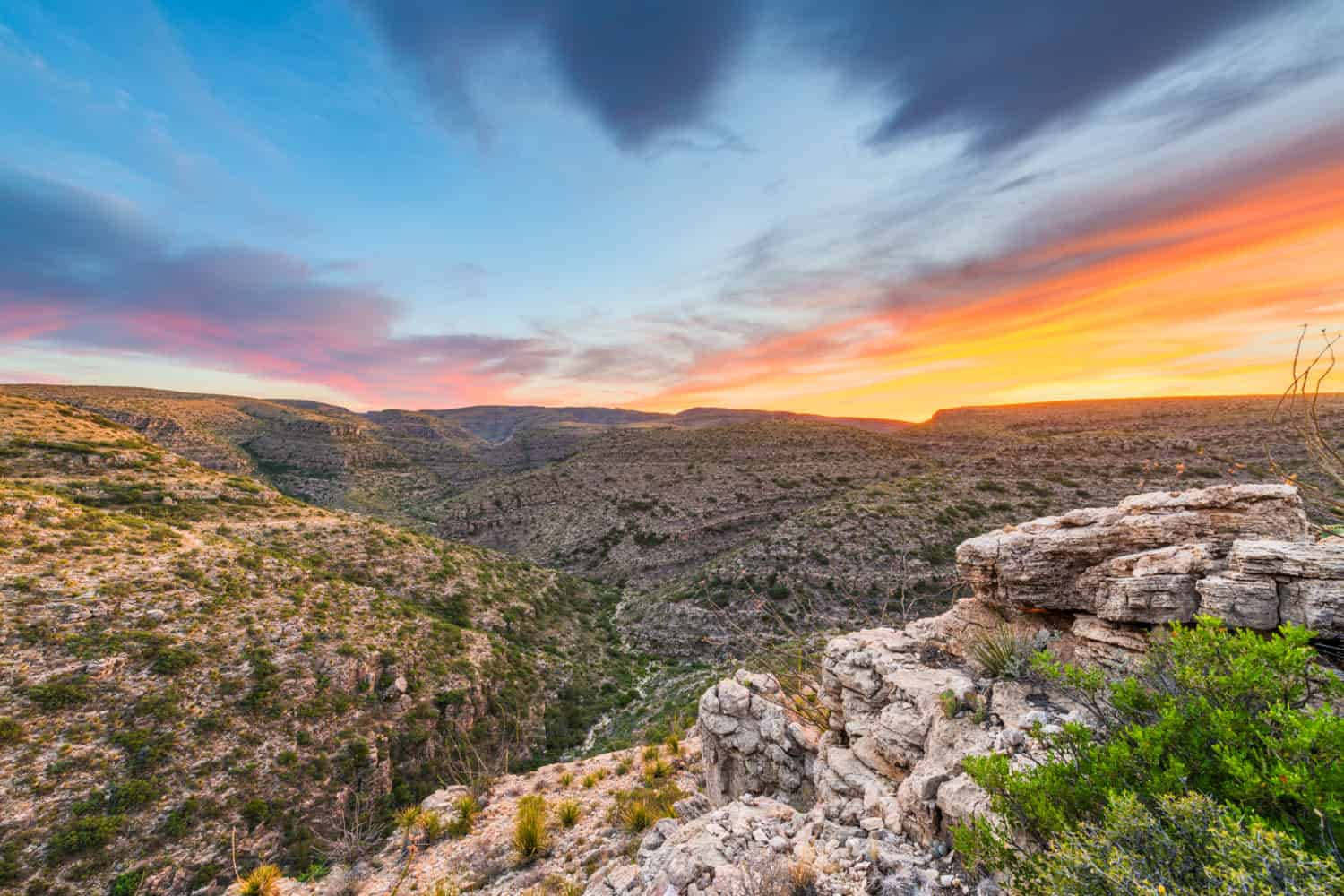 New Mexico 桌布 壁紙 — 令人嘆為觀止的日落將天空染成火熱的橙色和粉紅色，映照在崎嶇的峽谷景觀上，有岩石峭壁和稀疏的綠色植物。
