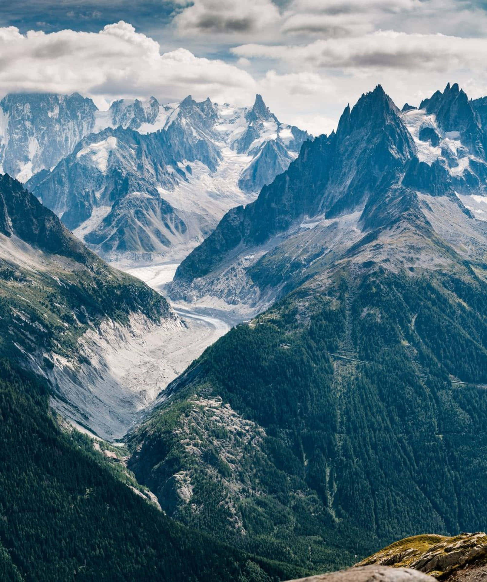 山景 桌布 壁紙 — 令人嘆為觀止的景色，白雪覆蓋的山峰和冰川蜿蜒穿過深邃的森林山谷，天空多雲。