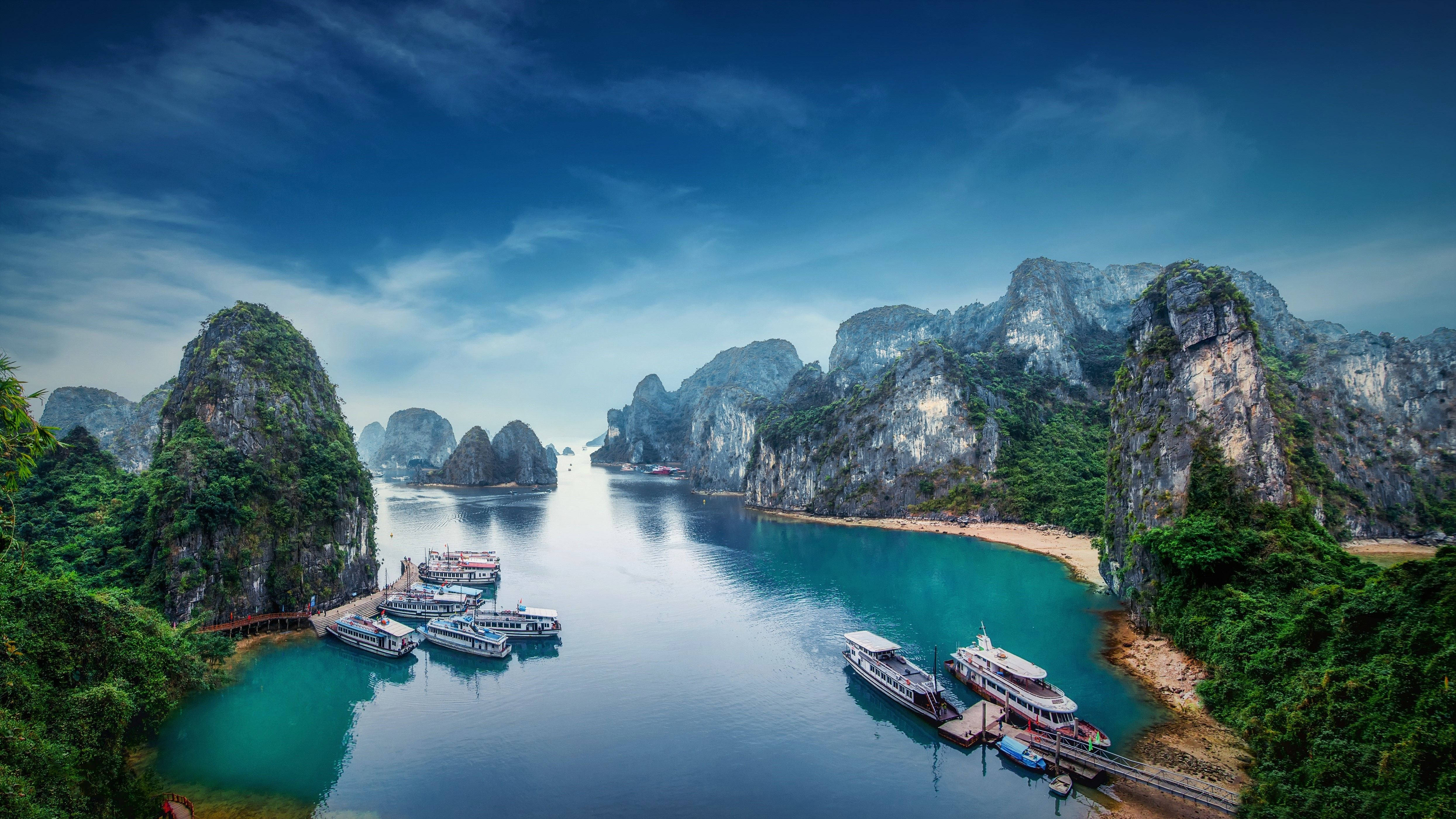 Hanoi 桌布 壁紙 — 令人嘆為觀止的景色，高聳的石灰岩喀斯特地貌從碧綠的海水中升起，船隻停泊在寧靜的海灣，上方是戲劇性的藍天。