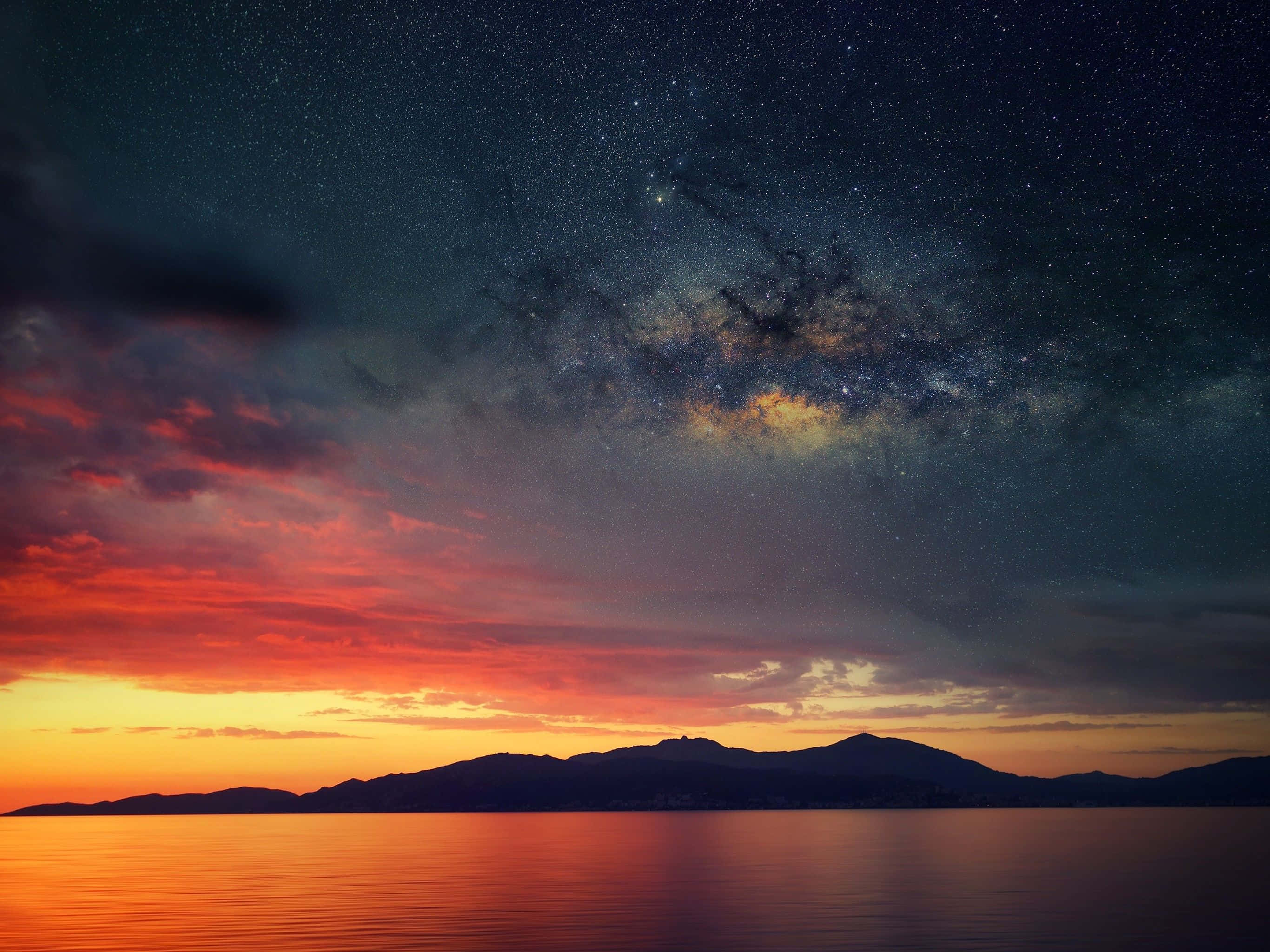 夜空 桌布 壁紙 — 令人嘆為觀止的暮色天空，火紅的橙色雲彩和生動的銀河拱跨在剪影般的山脈和寧靜的水面上。