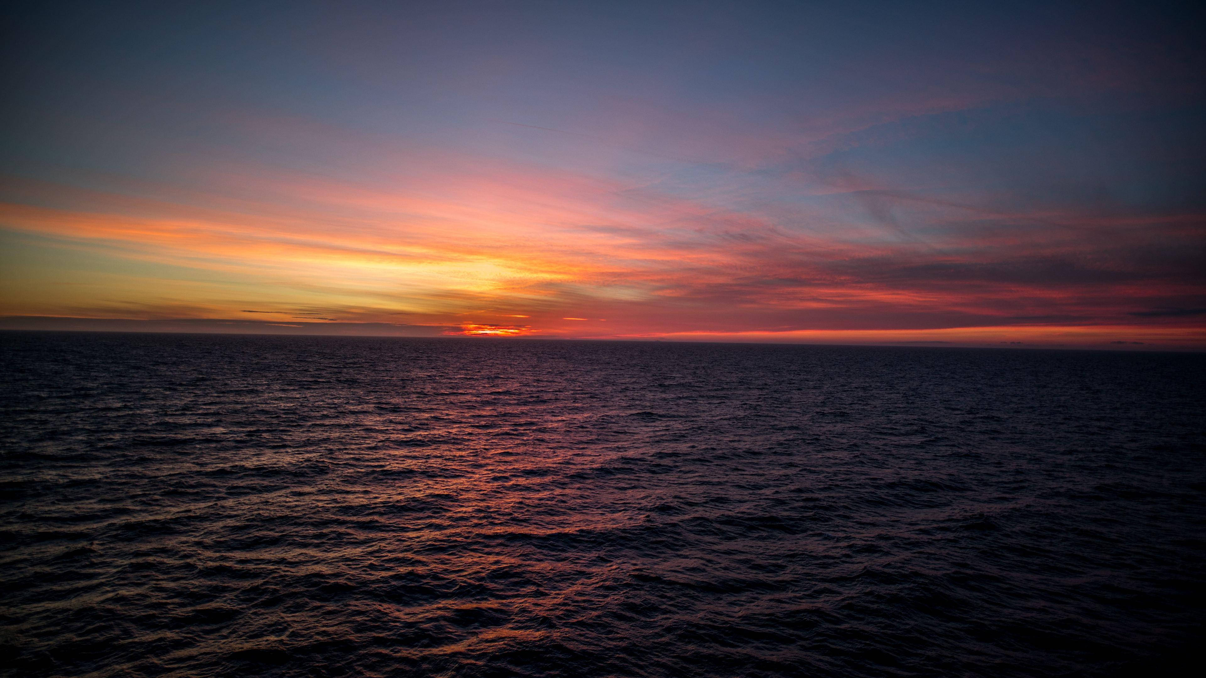 Sunset Aesthetic 桌布 壁紙 — 令人嘆為觀止的海洋日落，火紅的橙色和粉紅色雲彩倒映在黑暗的漣漪水面上。