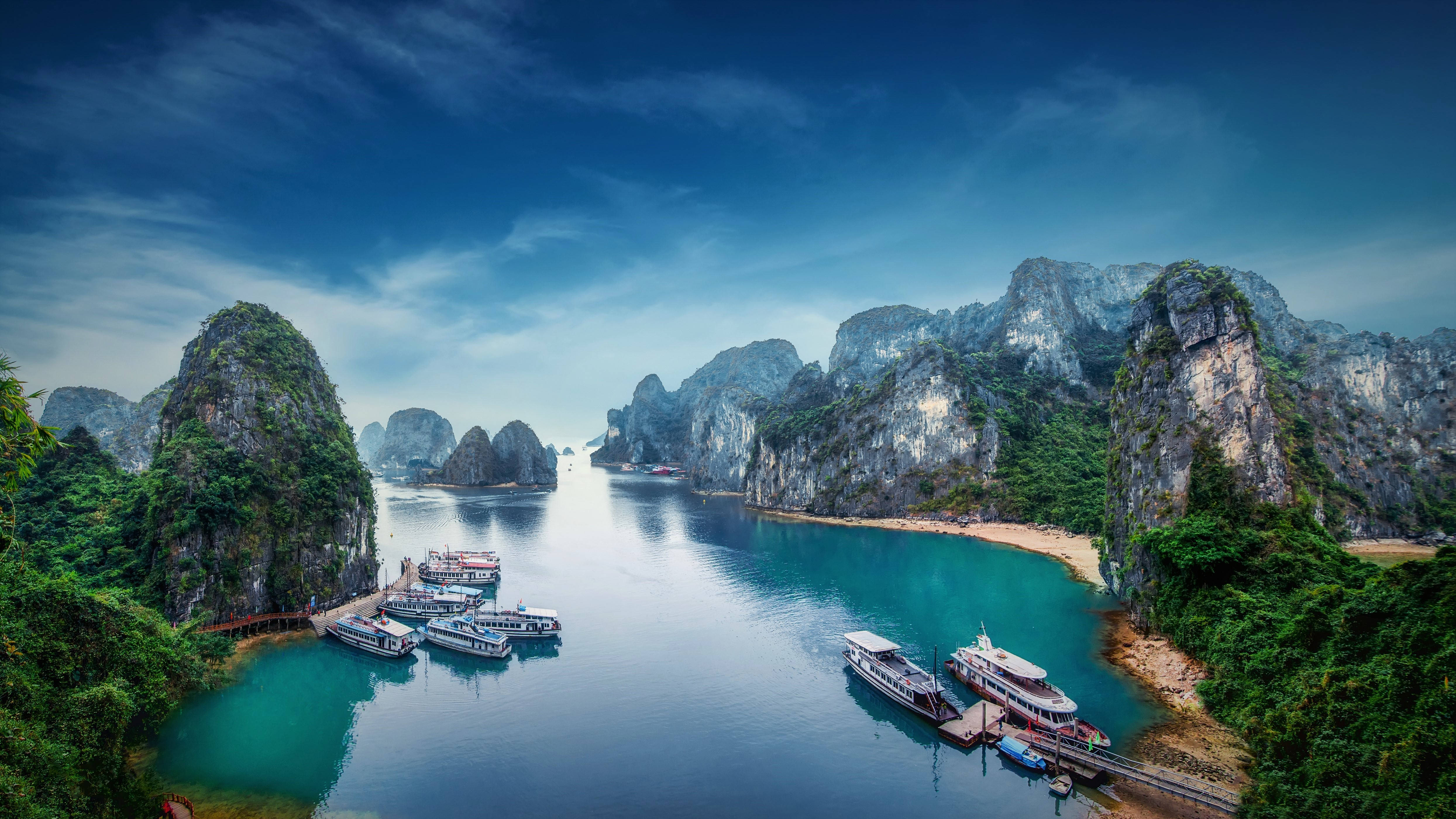 Vietnam 桌布 壁紙 — 令人嘆為觀止的鳥瞰圖，高聳的石灰岩喀斯特地貌覆蓋著茂密的綠色植物，從碧綠的海水中升起，點綴著停泊的船隻，在戲劇性的藍天下。