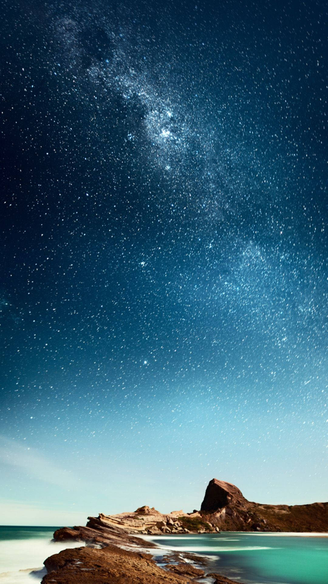 海灘 iPhone 桌布 壁紙 — 令人屏息的夜空，佈滿無數星辰，銀河拱跨在寧靜的碧綠海洋與崎嶇的海岸岩石之上。
