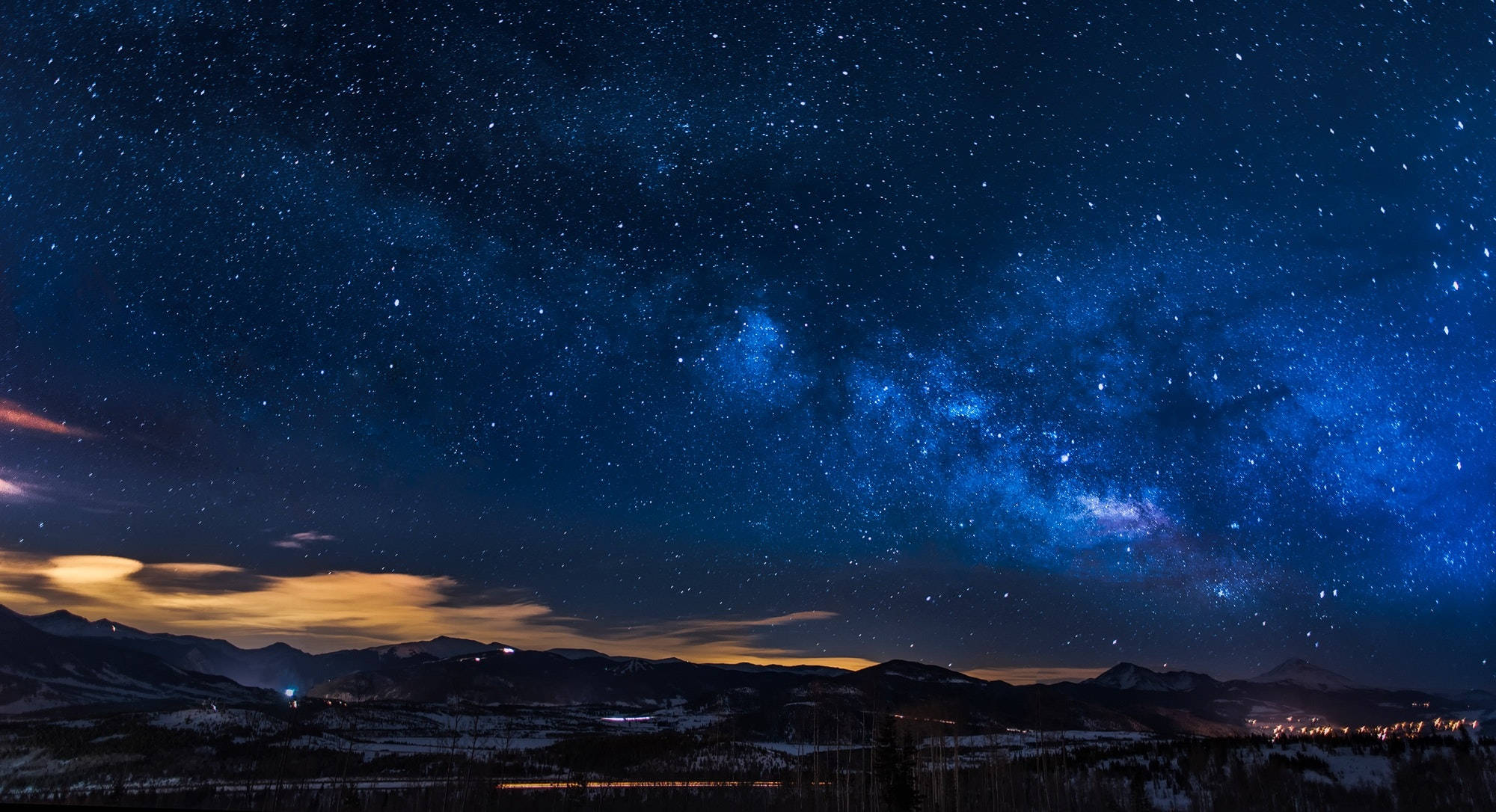 Stars 桌布 壁紙 — 令人屏息的夜空，充滿無數星星，閃爍的銀河拱跨在積雪的山脈之上，下方是遙遠的城市燈光。