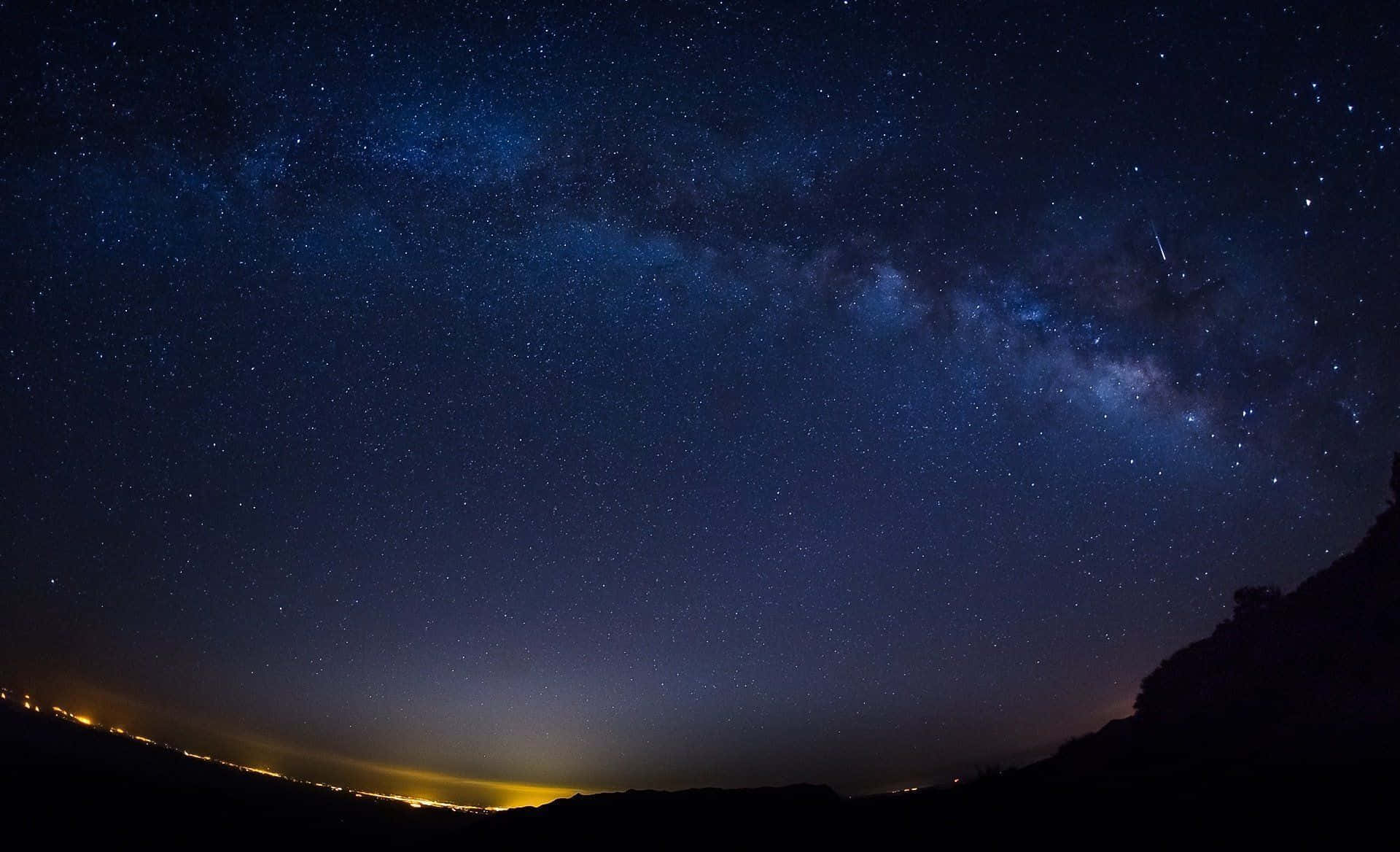 Starry Sky 桌布 壁紙 — 令人屏息的夜空，充滿無數星星和發光的銀河，在黑暗的山丘輪廓上拱起，下方是遠處的城市燈光。
