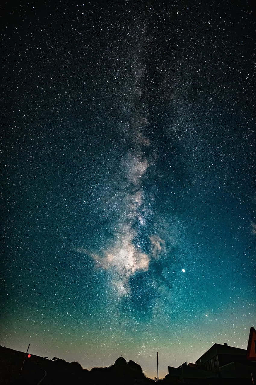 Astrophysics 桌布 壁紙 — 令人屏息的夜空，充滿無數星星和閃爍的銀河，在屋頂和山丘的黑暗剪影上拱起。