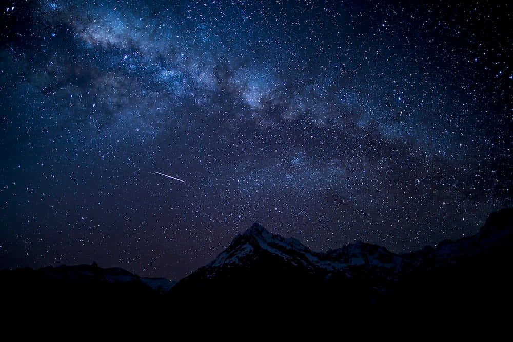 Night Time 桌布 壁紙 — 令人屏息的夜空，滿天繁星和銀河拱起在積雪的山峰之上，一顆流星劃過天際。