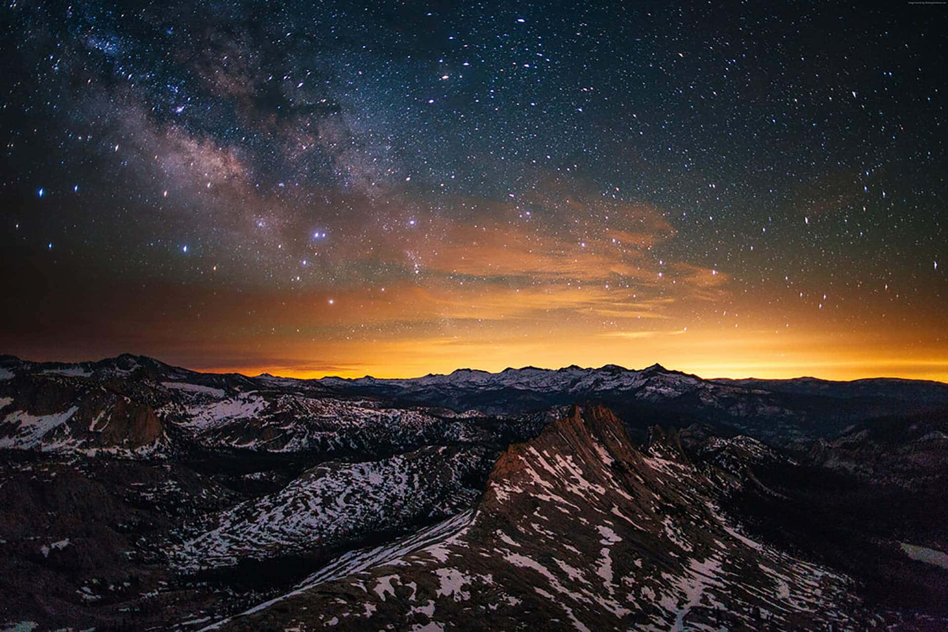 銀河 桌布 壁紙 — 令人屏息的夜空佈滿繁星，銀河在白雪覆蓋的山峰上拱起，下方是發光的橙色地平線。